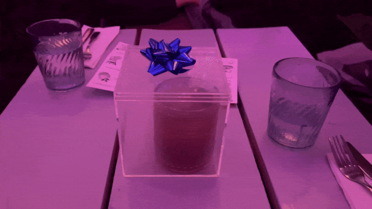 A GIF of a translucent box with a blue bow on top and a cocktail inside. A hand comes into the frame to remove the box's lid, unleashing a plume of smoke.