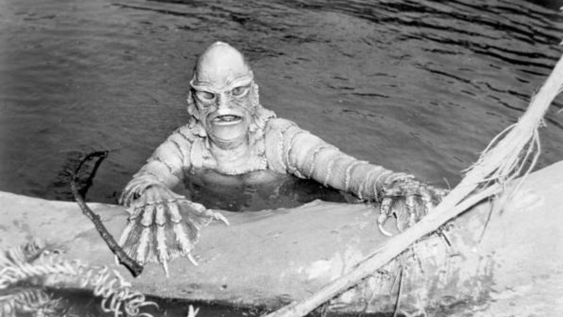 A black-and-white photo of a person dressed in a scaly monster costume holding onto a log with skeletal-like claws.