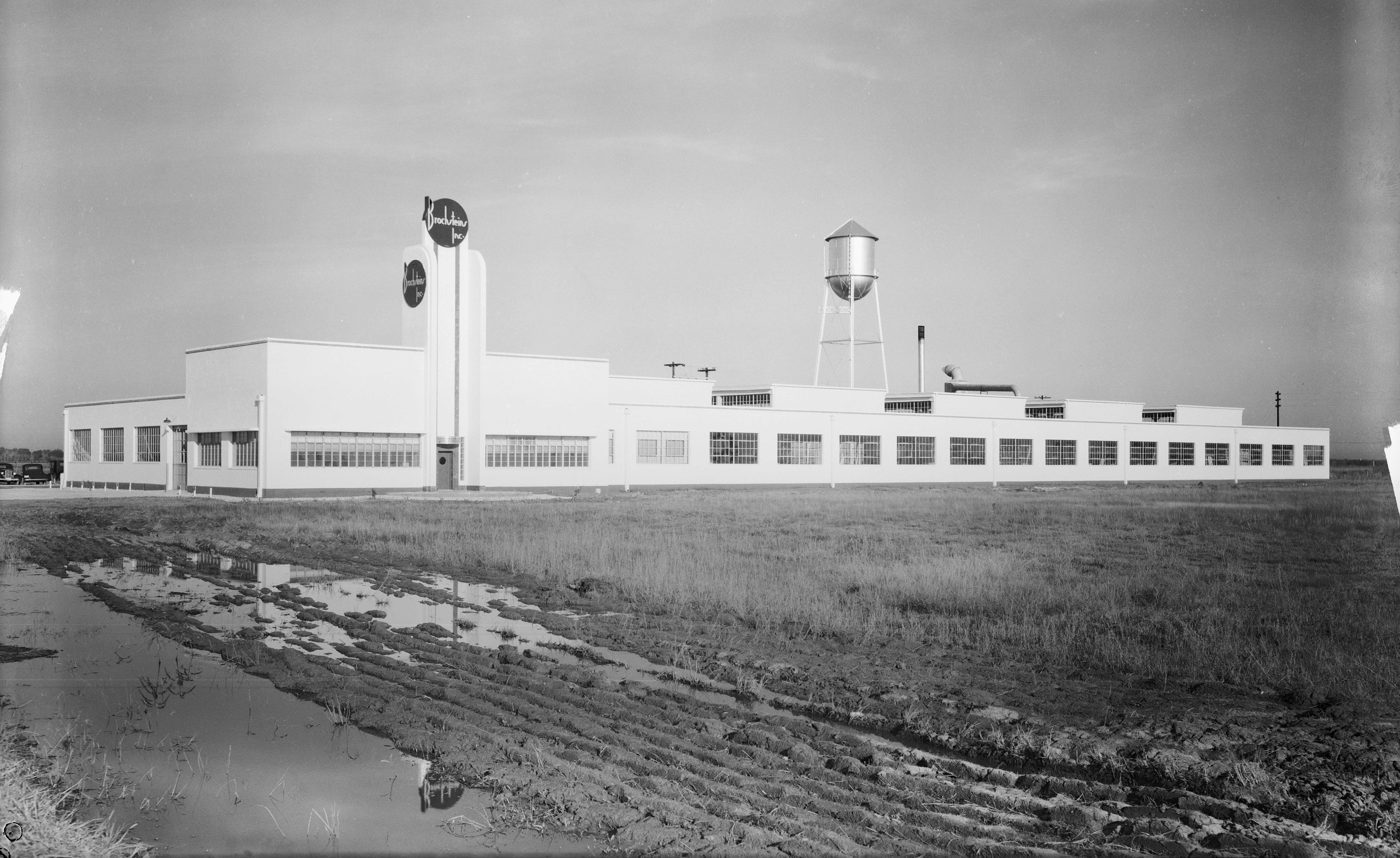Black-and-white photo of a long white industrial building with many windows, a tall water tower, and a grassy, muddy foreground; a vertical sign with circular logos stands at the front.