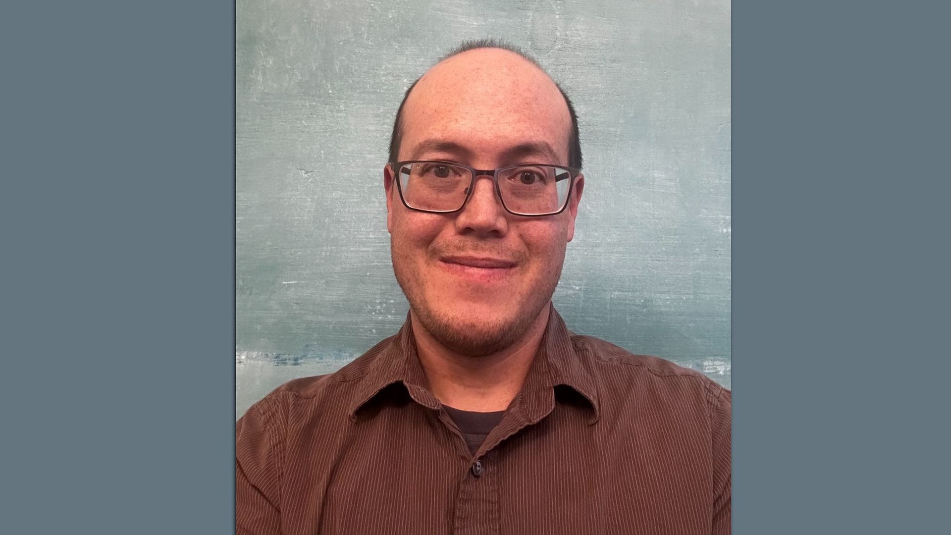 Smiling man with glasses, short hair, and a light beard wearing a brown shirt against a textured blue-gray wall background.