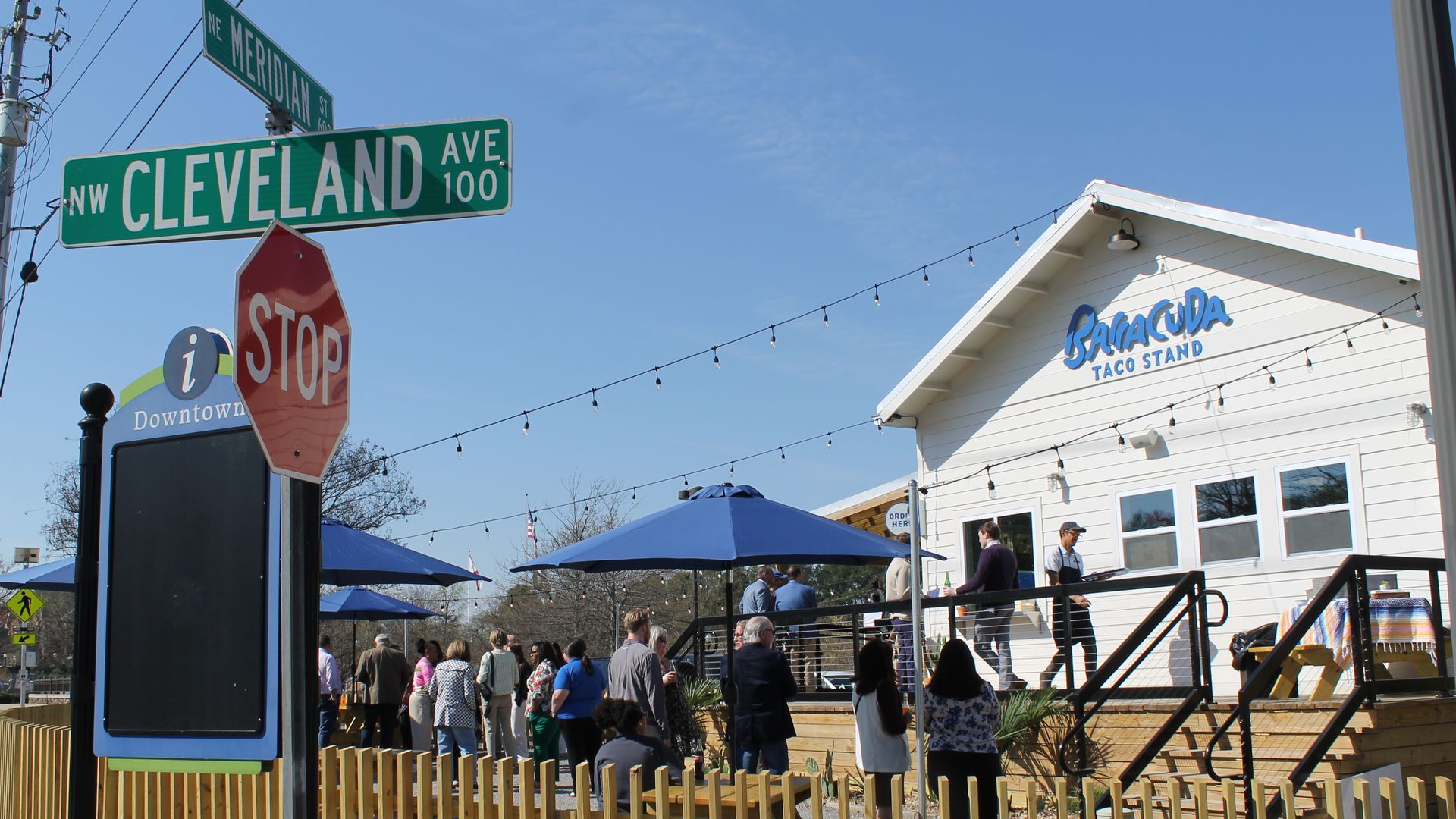 Sunny street corner with green Cleveland Ave and Meridian St signs, red stop sign, crowd gathered outside Batacuda Taco Stand under string lights and blue umbrellas; wooden fence encloses outdoor seating.