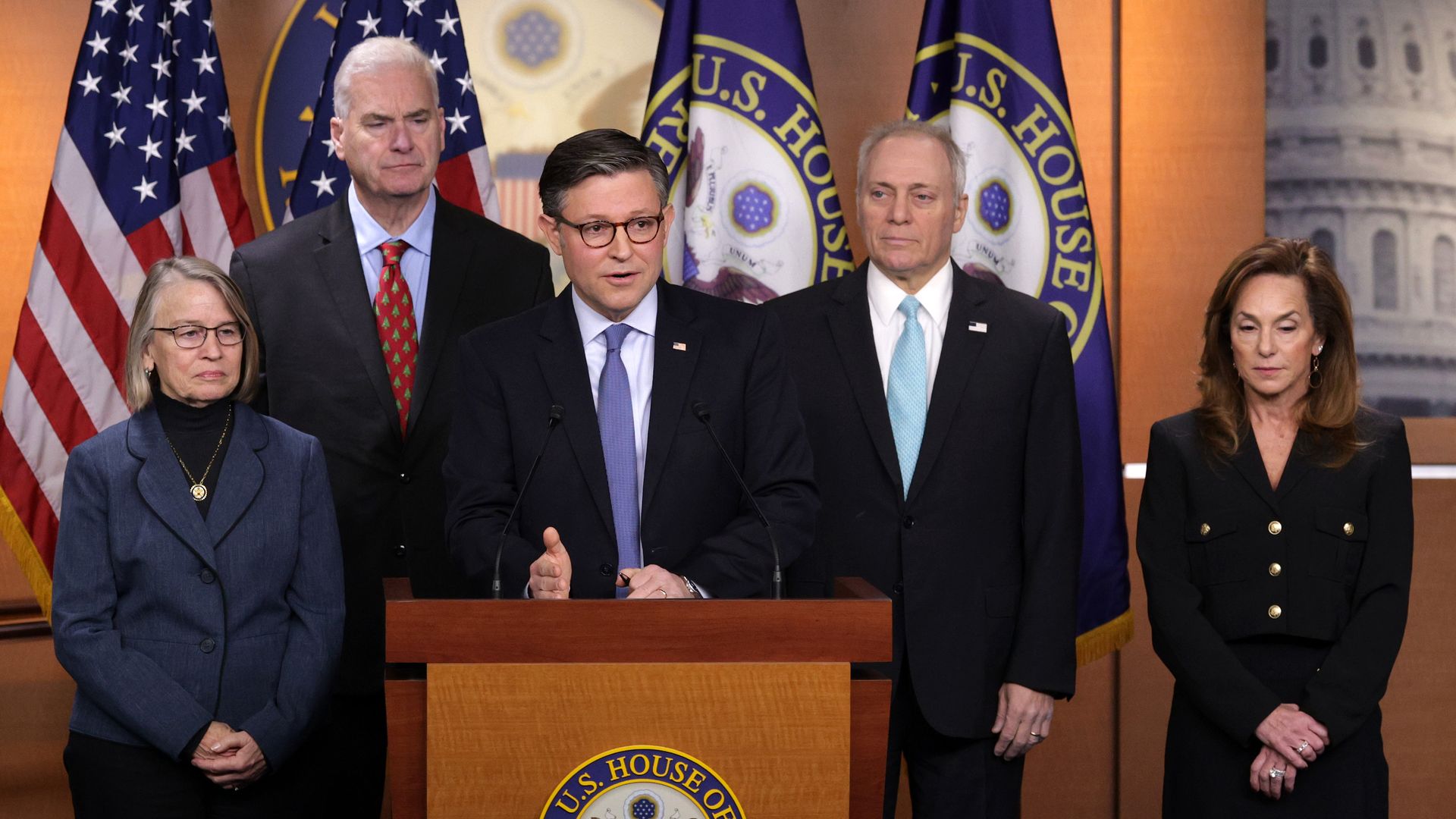 Five people in business attire stand behind a podium bearing the U.S. House seal, with American flags and blue banners framing the scene.