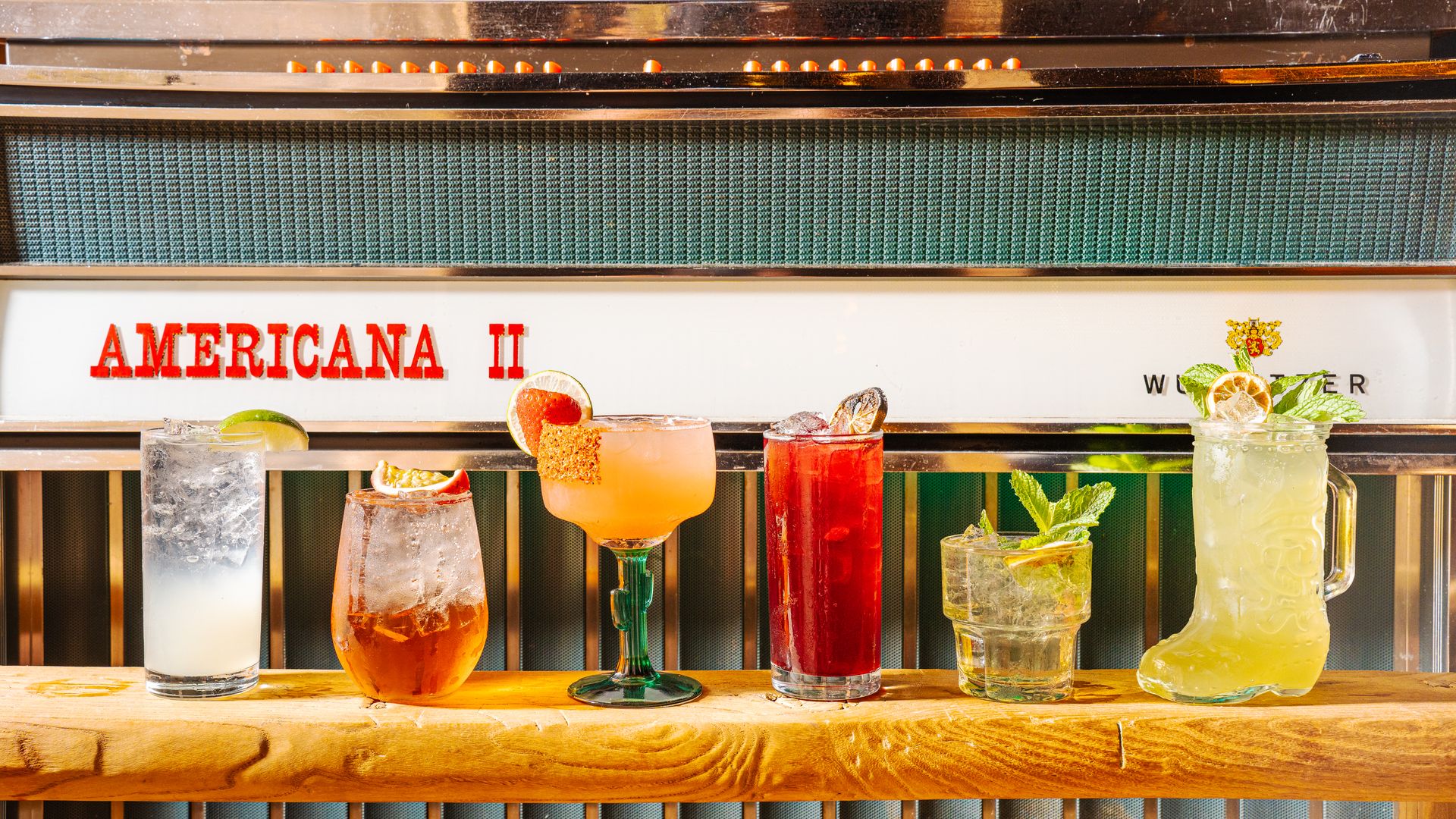Six colorful cocktails lined up on a wooden bar in front of a vintage jukebox labeled "AMERICANA II," featuring garnishes like lime, mint, and fruit slices.