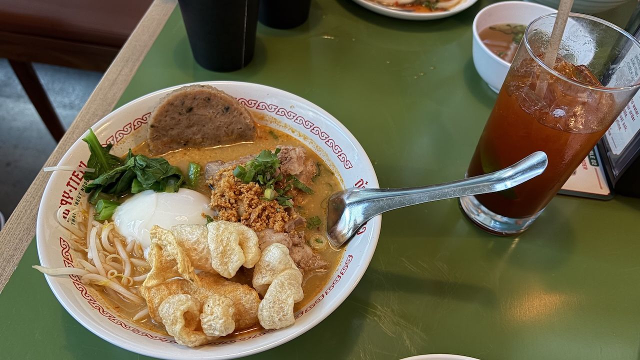 Top-down view of a ramen bowl with a soft-boiled egg, greens, bean sprouts, fried garlic, and pork crackling in a creamy broth; a glass of iced tea sits on a green table beside the bowl.
