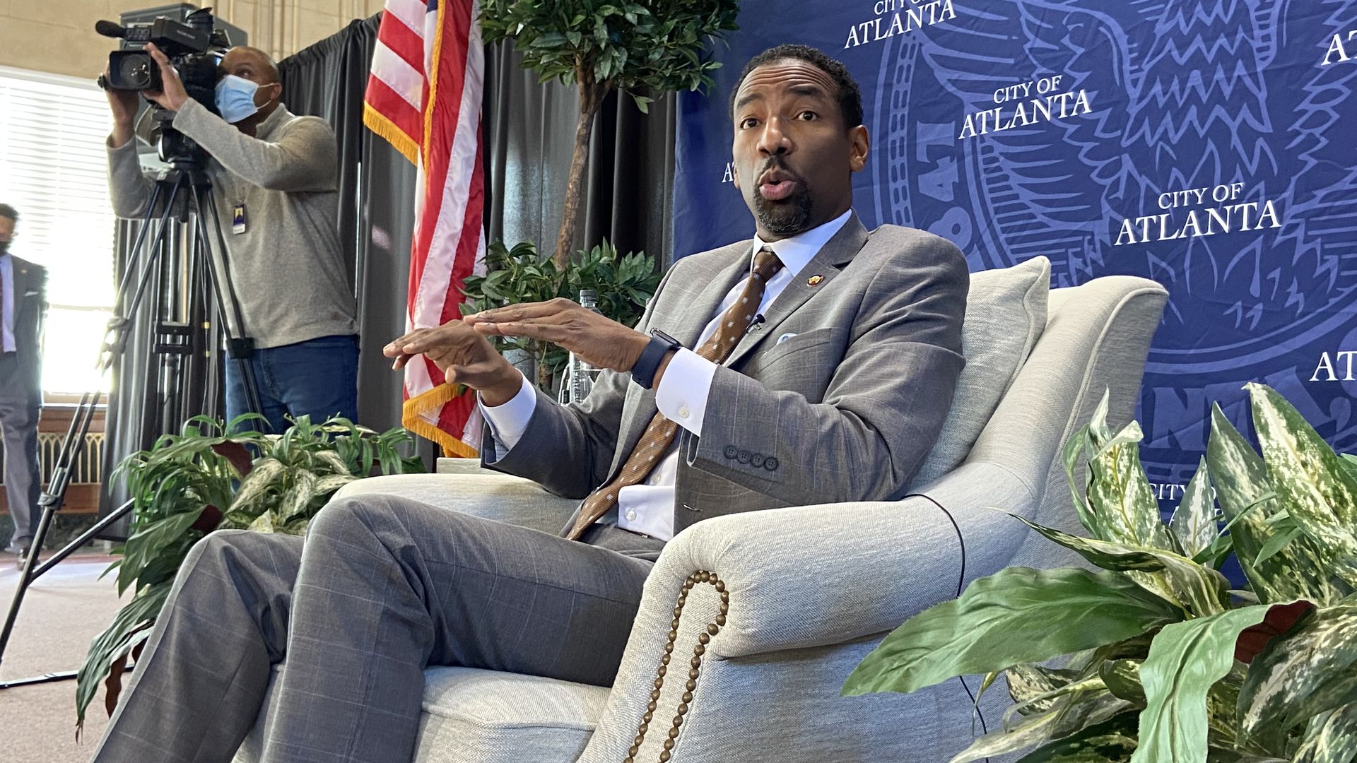 Mayor Andre Dickens, wearing a gray suit, sits in a white chair in front of blue backdrop that says "City of Atlanta"
