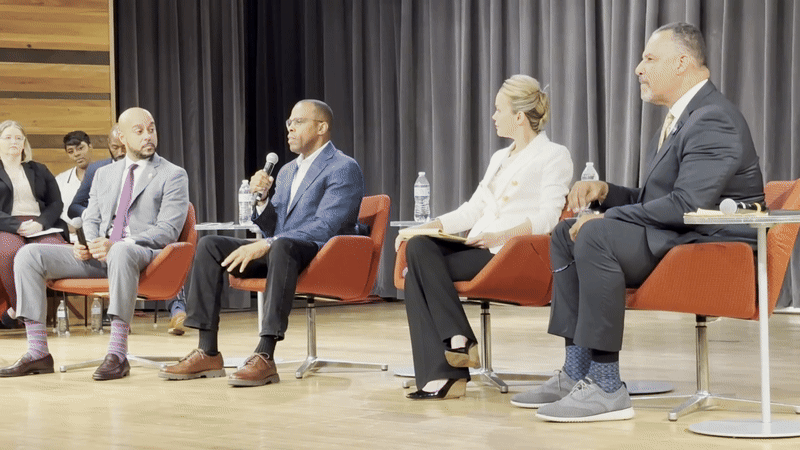 Five panelists seated on stage in red chairs during a discussion, with one man speaking into a microphone, a gray curtain backdrop, and two water bottles on small round tables.
