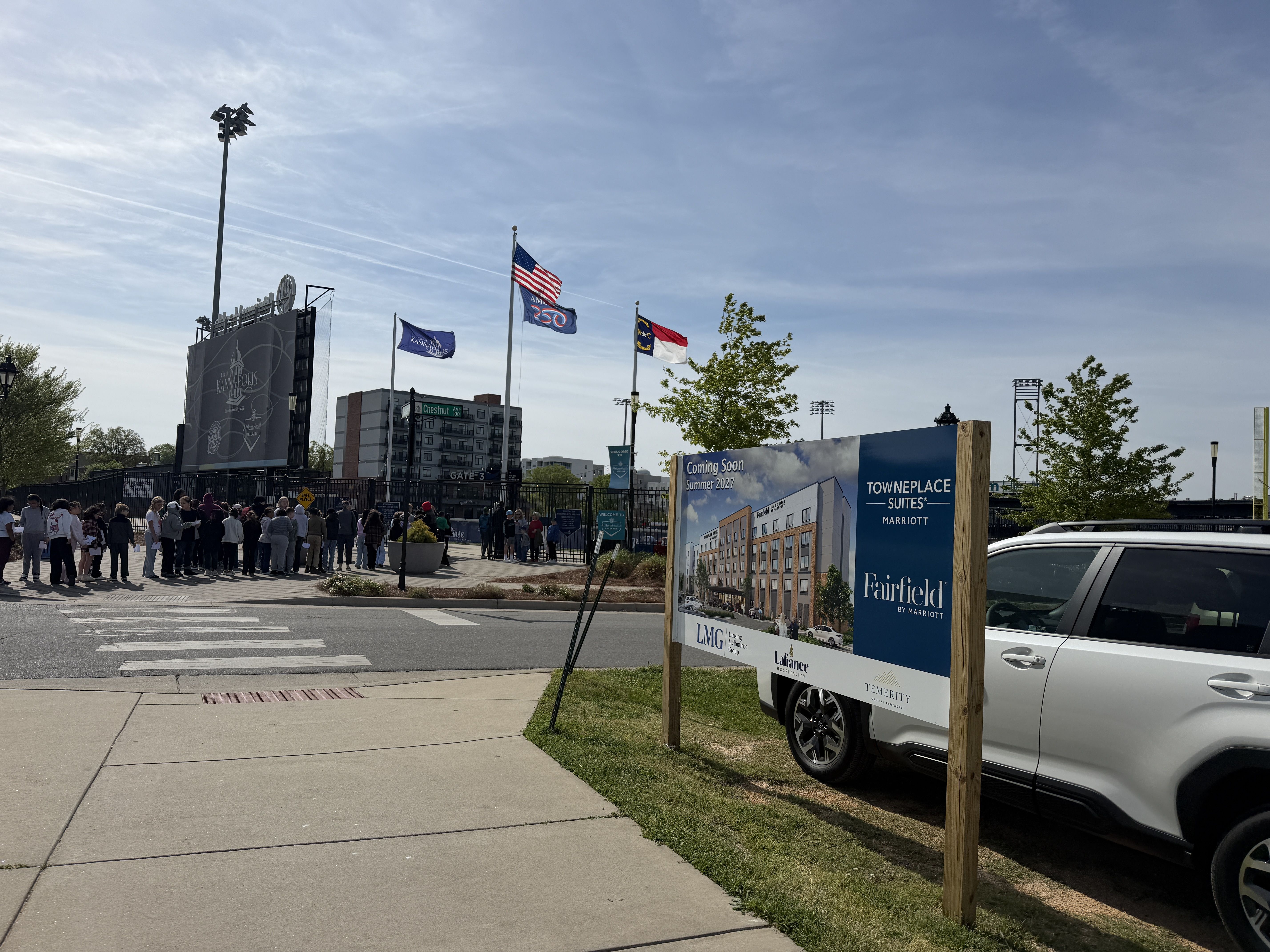 Wide view of a stadium entrance with a crowd lined up by a crosswalk, a tall scoreboard, flagpoles with the American flag, and a large blue sign near a white SUV.