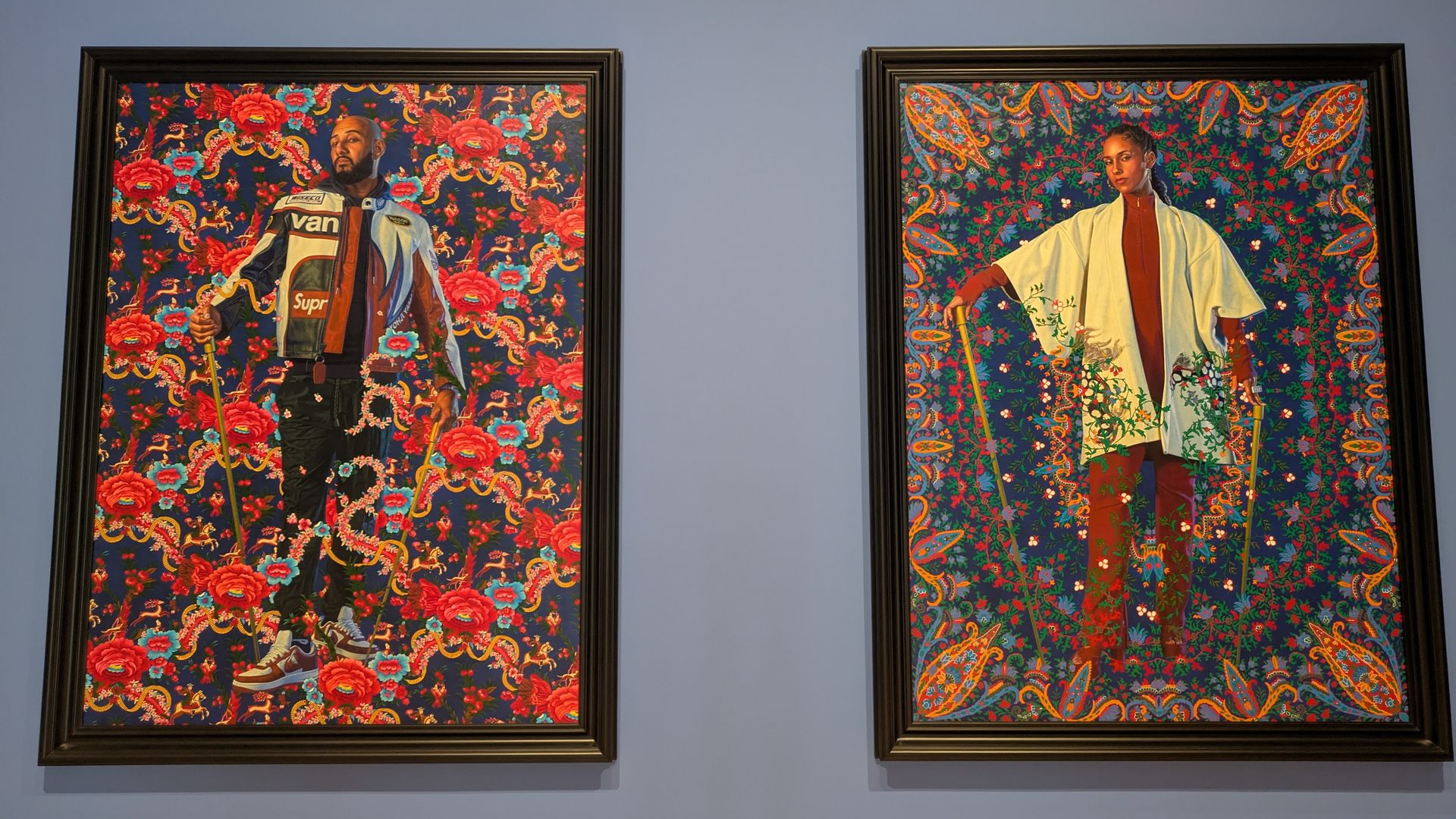 Two framed portraits on a blue wall: left, a bearded man in a colorful racing jacket before a red floral backdrop; right, a woman in a pale robe before a vibrant, patterned background.
