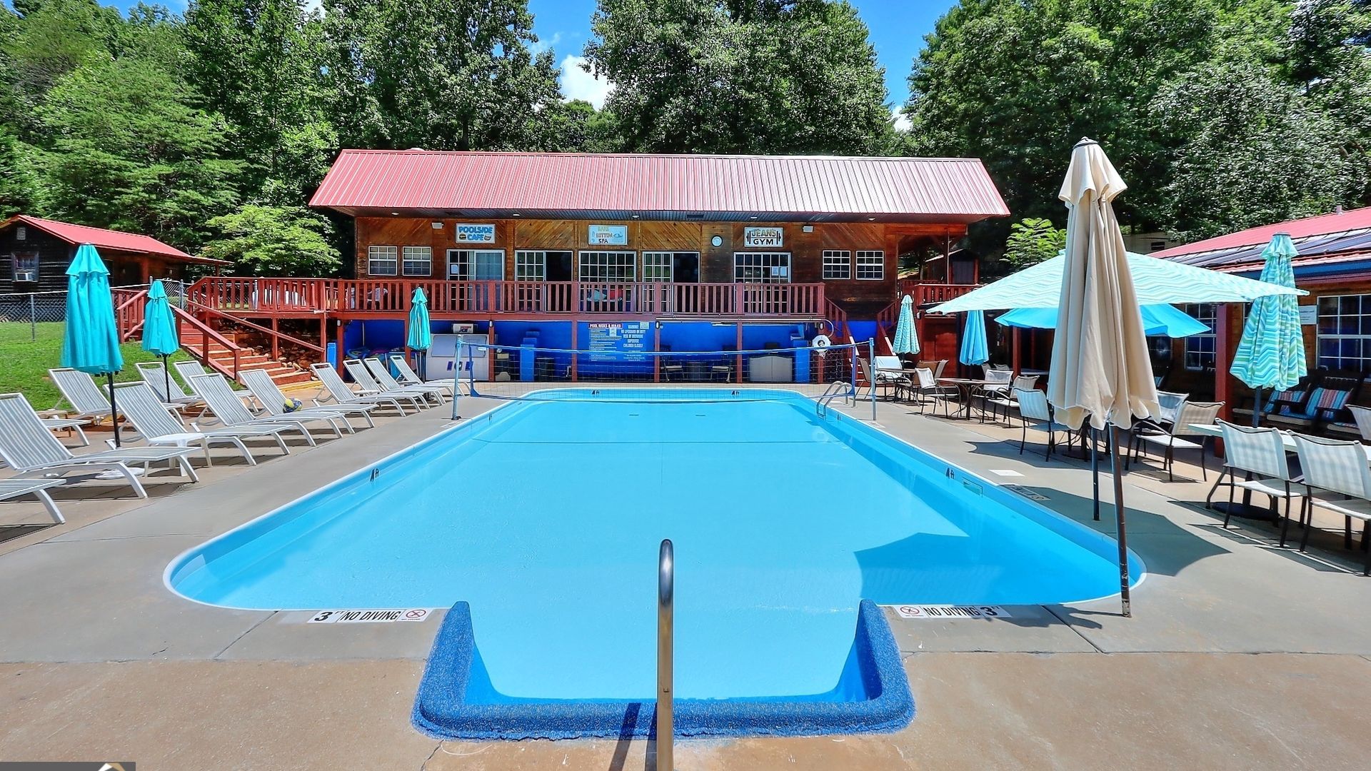 A photograph of a blue swimming pool surrounded by folded-down umbrellas and lounge chairs. The pool abutted by a pavilion and a clubhouse