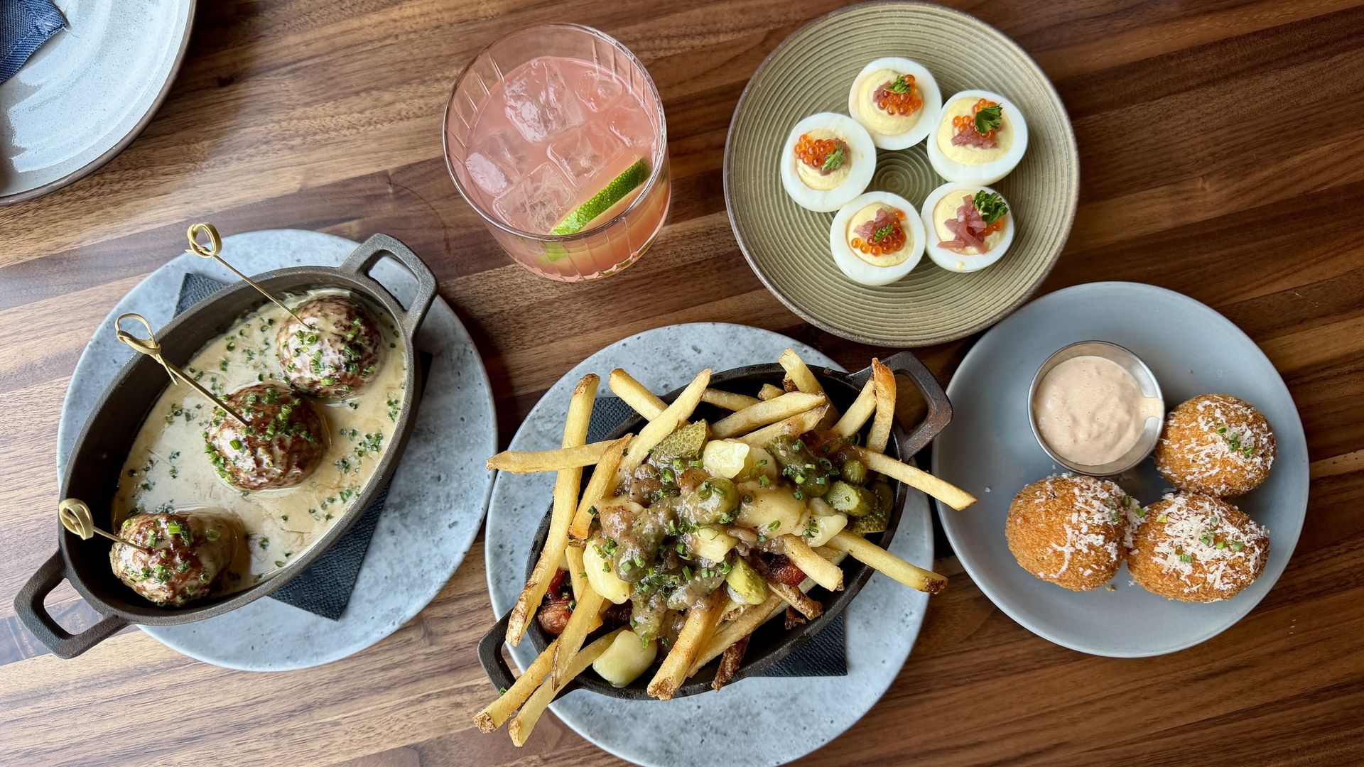 Wood table with several dishes: meatballs in creamy sauce in a skillet, a plate of loaded fries with toppings, deviled eggs with caviar, three fried arancini with dip, and a pink cocktail with lime.