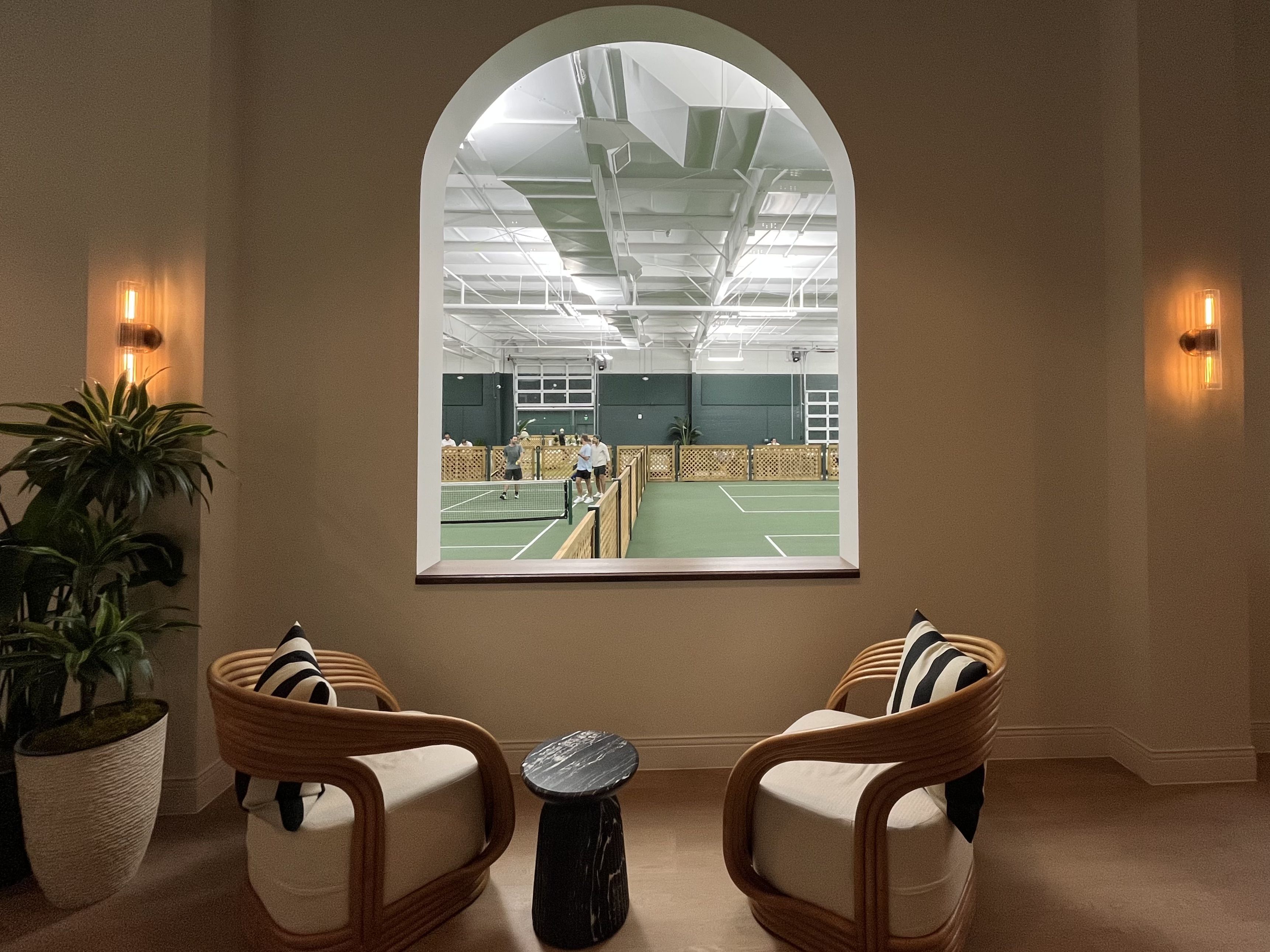 Interior view of a cozy beige seating area with two wooden armchairs and a small black table, looking through an arched window to an indoor pickleball court where people play on green courts.