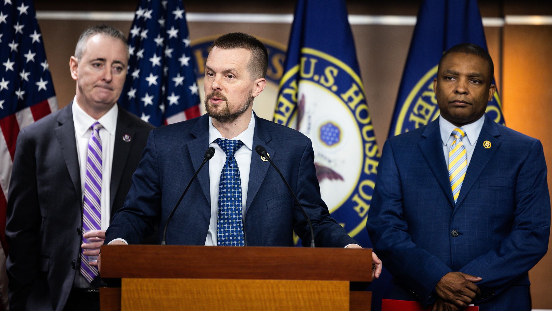 Rep. Jared Golden, wearing a blue suit, white shirt and blue tie, stands at a podium flanked by Reps. Brian Fitzpatrick, in a gray suit, and Don Davis, in a blue suit, in front of American and U.S. House of Representatives flags.