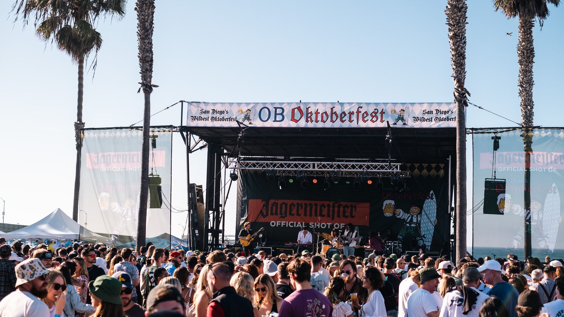 Outdoor Oktoberfest event in San Diego with a large crowd enjoying live music on stage under a banner reading "OB Oktoberfest," palm trees, clear blue sky, and ocean in the background.