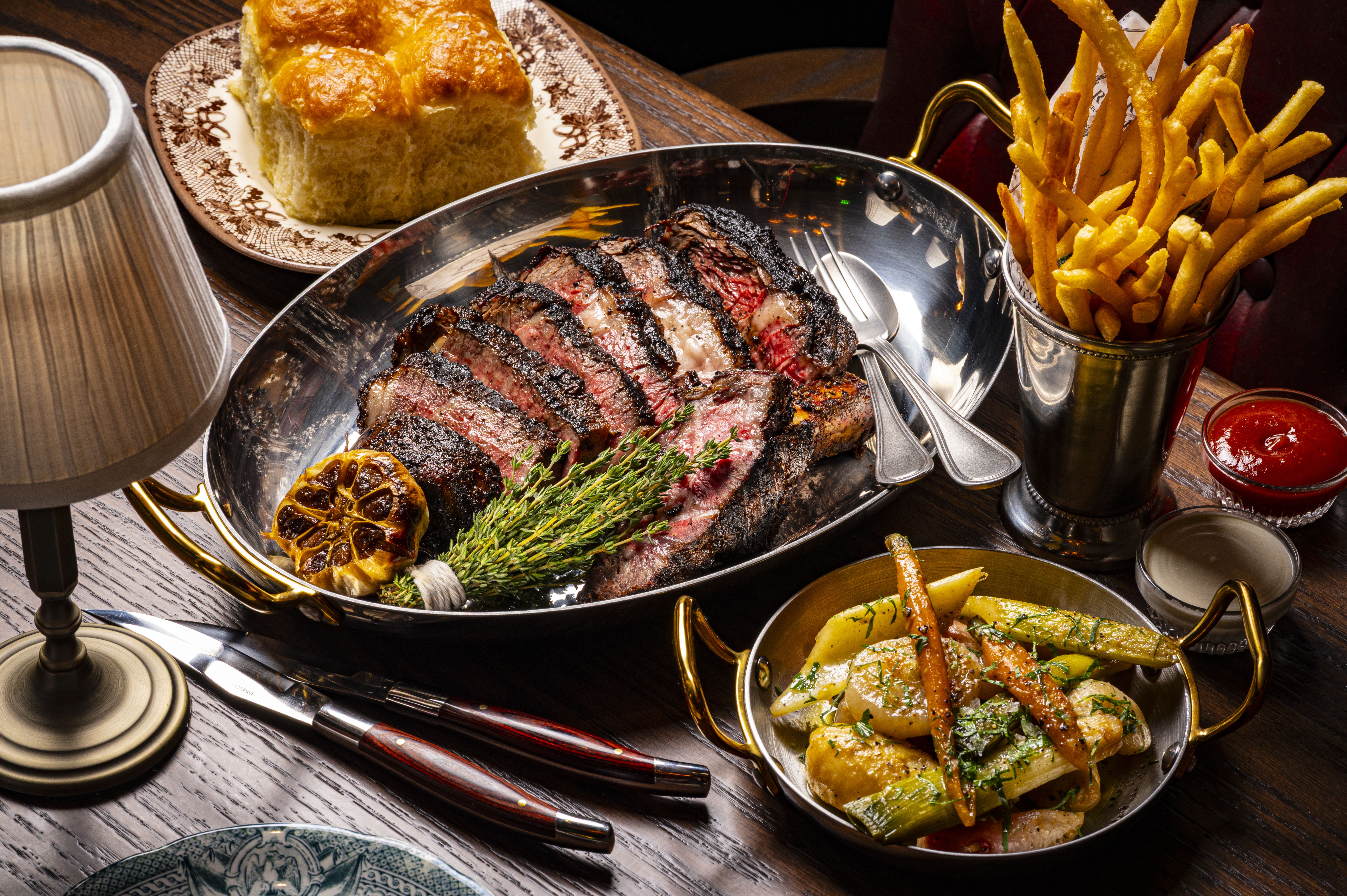 Sliced, charred steak in a shiny skillet with rosemary and lemon, surrounded by fries in a metal cup and a decorative bread loaf on a plate.