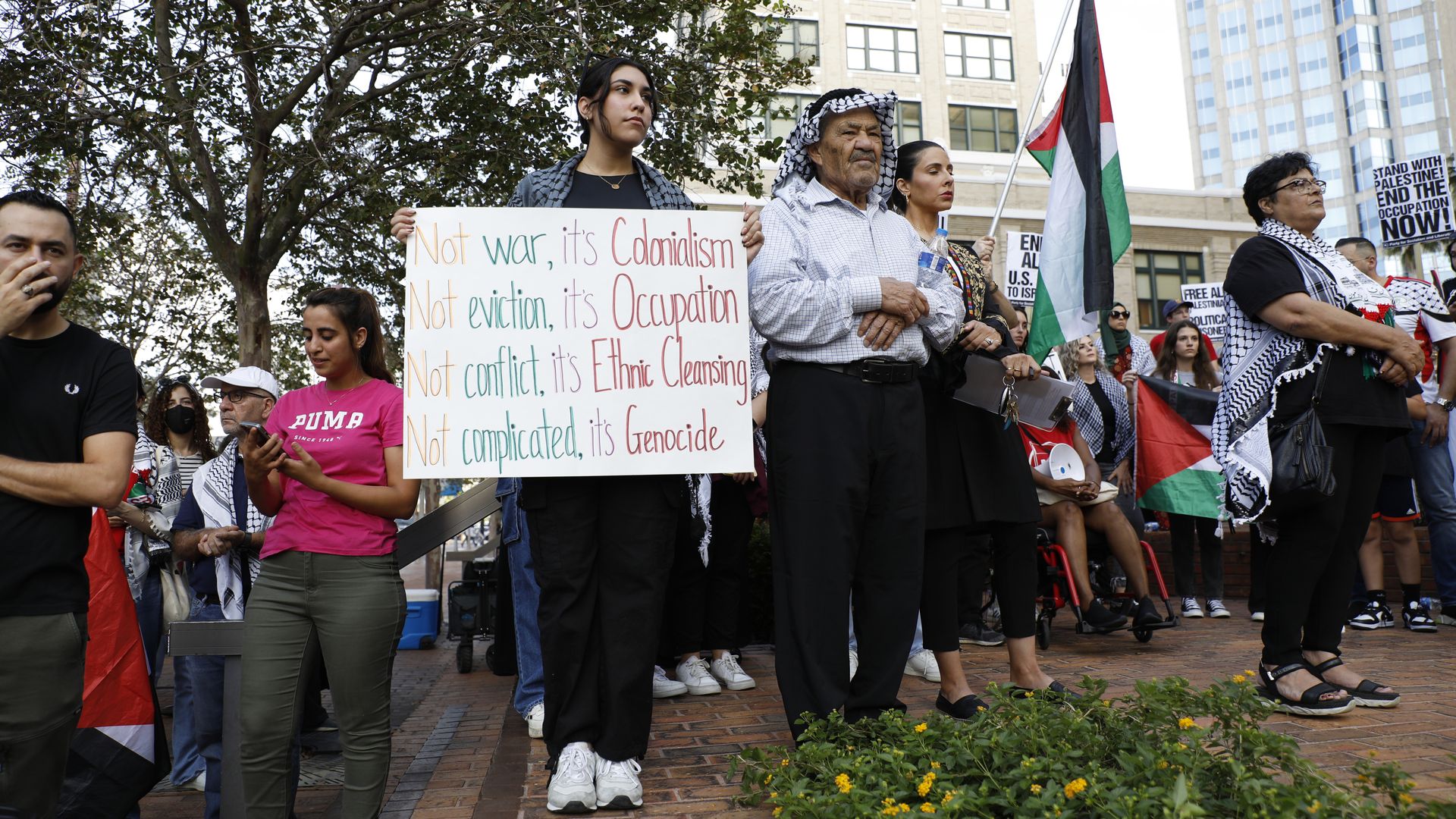 Demonstrators at a rally for Palestinians, including a woman holding a sign that says, "Not war, it's colonialism. Not eviction, it's occupation. Not conflict, it's ethnic cleansing. Not complicated, it's genocide."