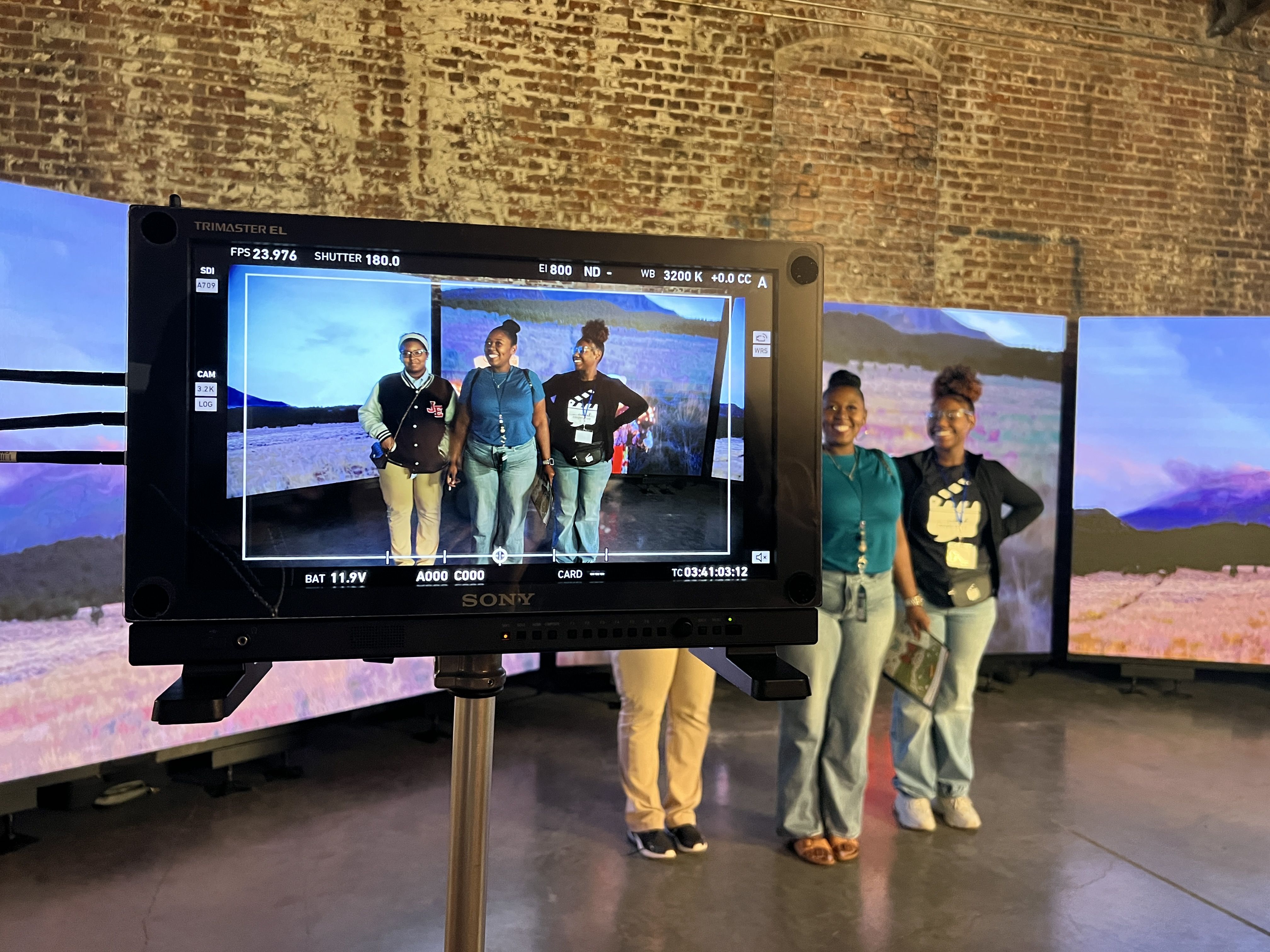 Three people standing in front of curved digital screens showing a scenic landscape are captured on a Sony camera monitor inside a room with brick walls and a concrete floor.