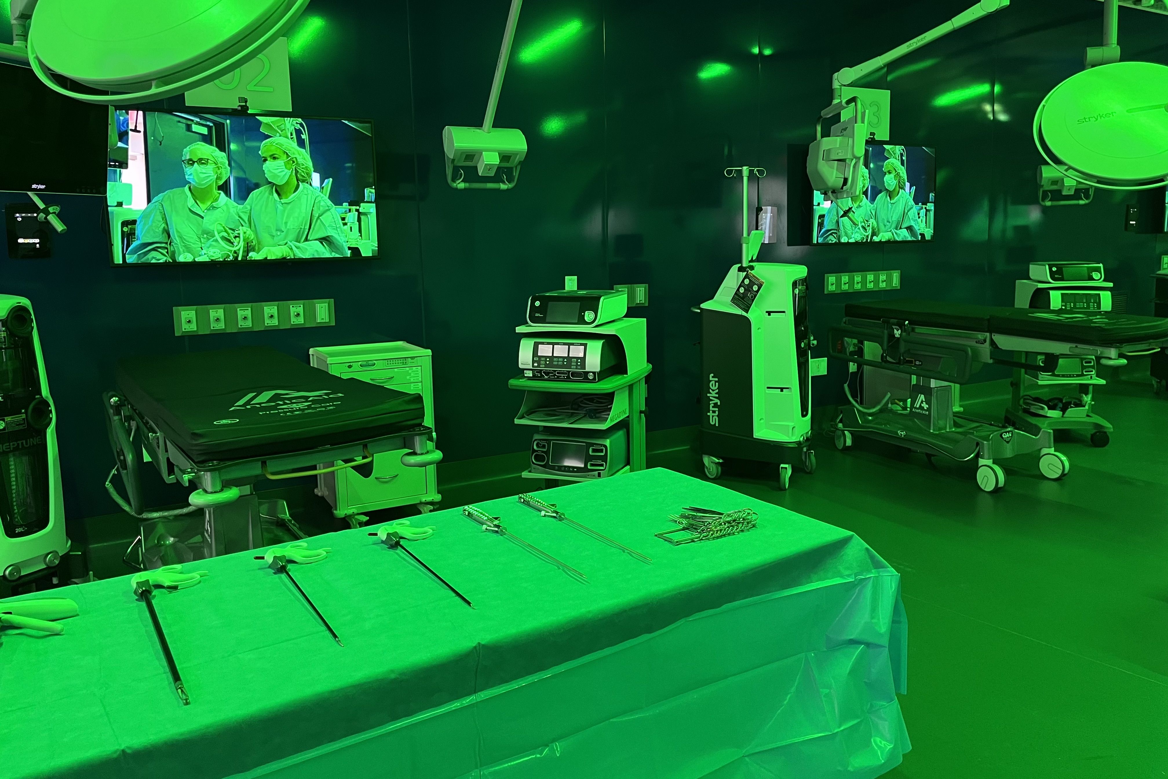 Operating room with surgical instruments on a table, medical monitors displaying healthcare workers in scrubs and masks, hospital beds, and advanced medical equipment under green lighting.