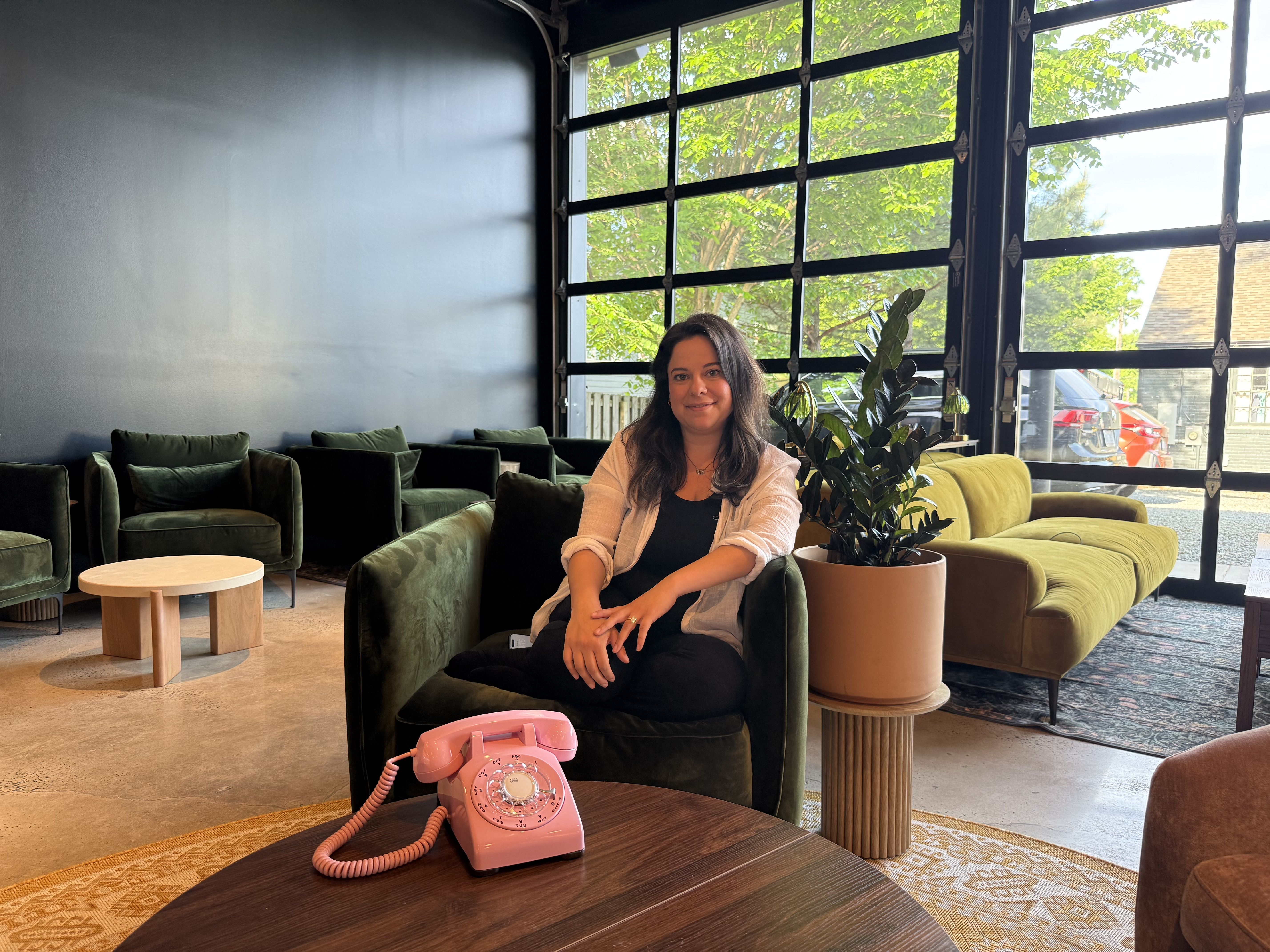 Woman seated in a dark green velvet chair inside a bright lounge; a pink rotary phone on a round table, a potted plant on a pedestal, and a yellow sofa by large glass doors showing trees outside.plot twist Nikki 