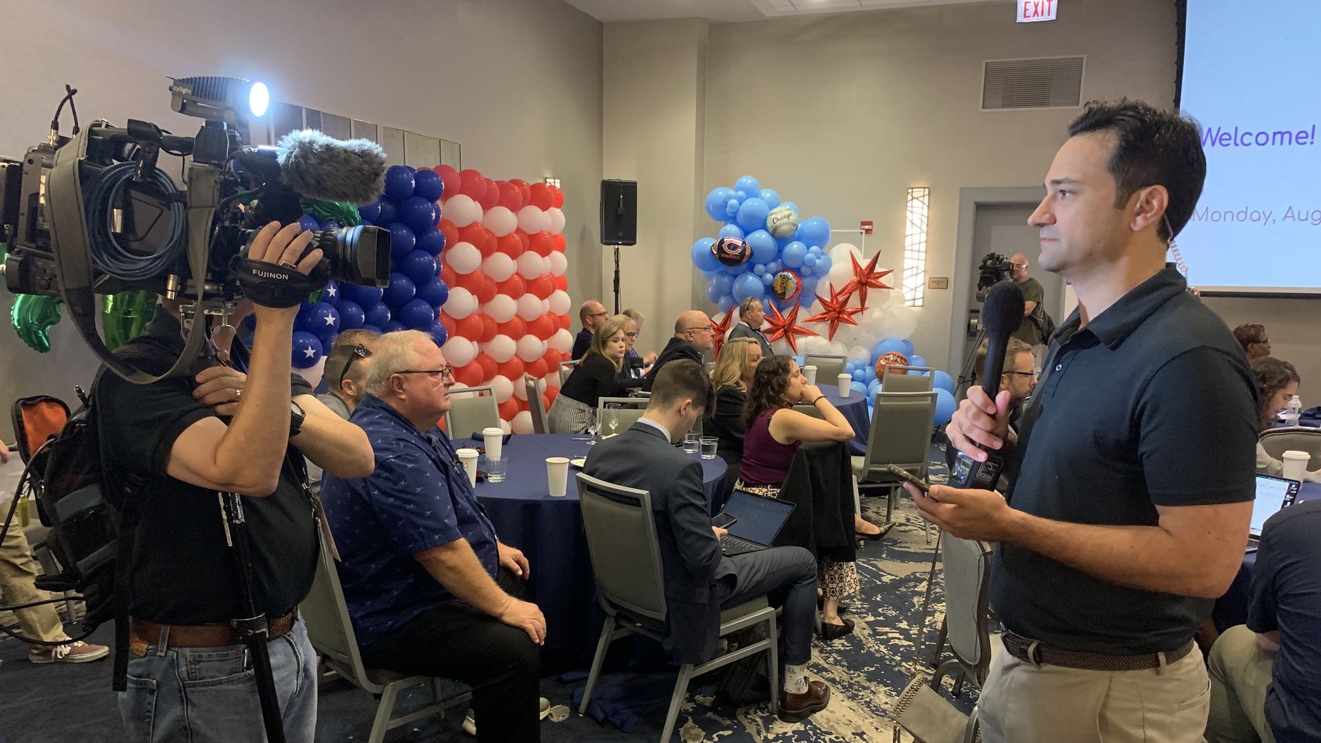 A cameraman with a large broadcast camera films a man holding a microphone at a conference. Red, white, and blue balloon arches decorate the room as attendees sit at tables and a "Welcome" slide appears on a screen.