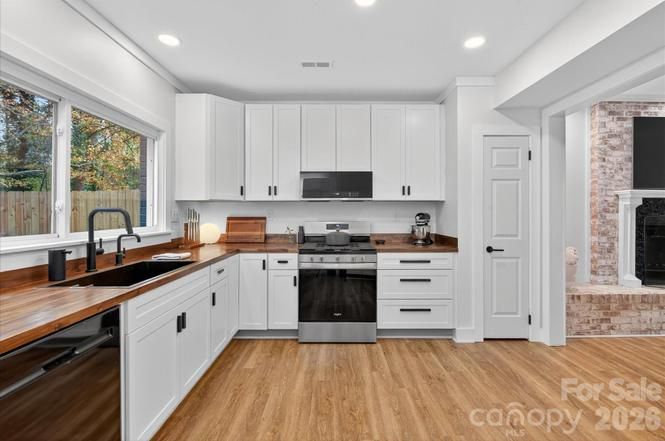 Modern kitchen with white cabinets, black hardware, wooden countertops, black sink and faucet, stainless steel stove, dishwasher, and large window overlooking a fenced yard.