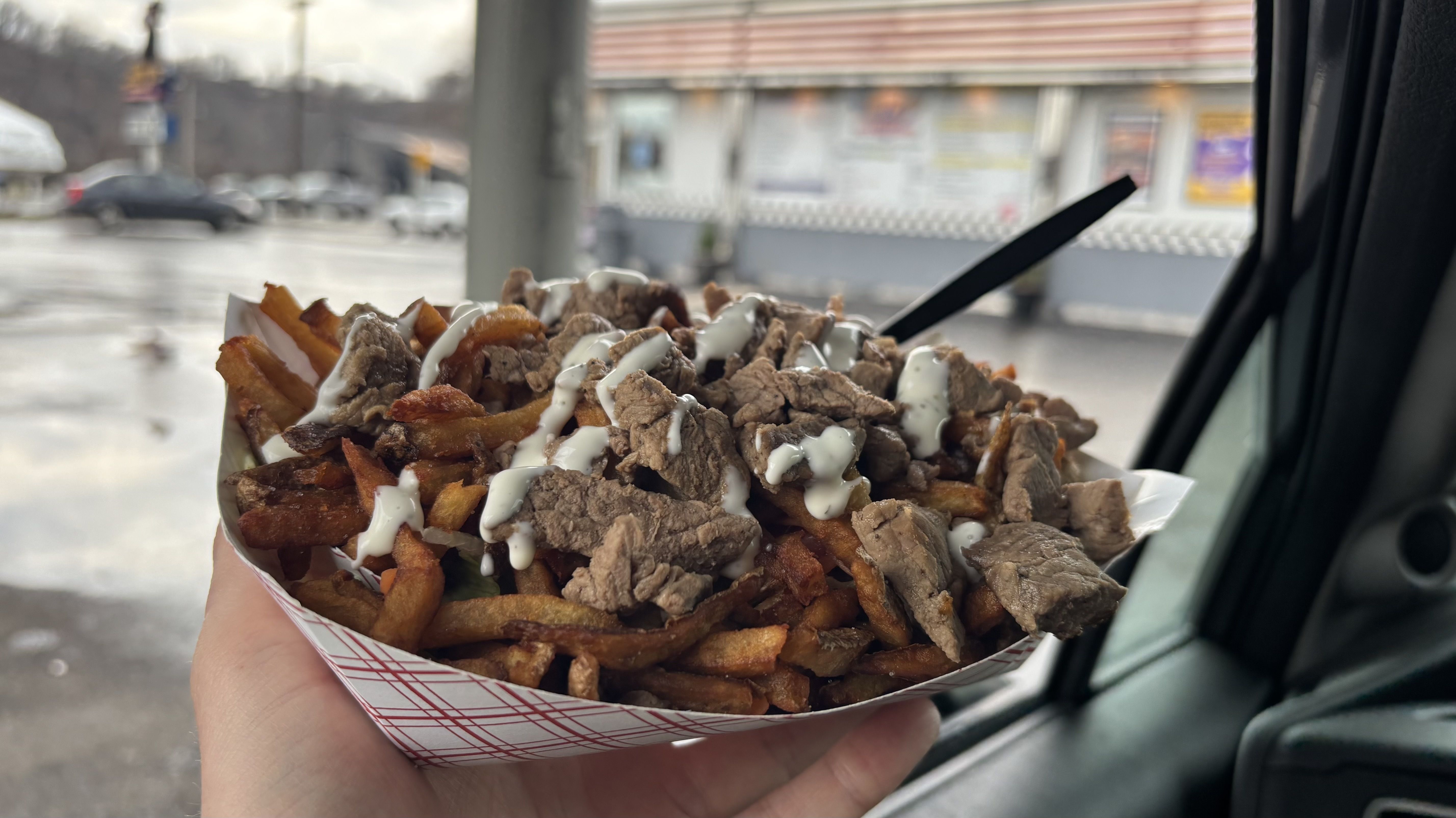 Hand holds a paper tray of loaded fries topped with beef chunks and a white sauce drizzle, inside a car with a blurred urban street and storefront in the background.