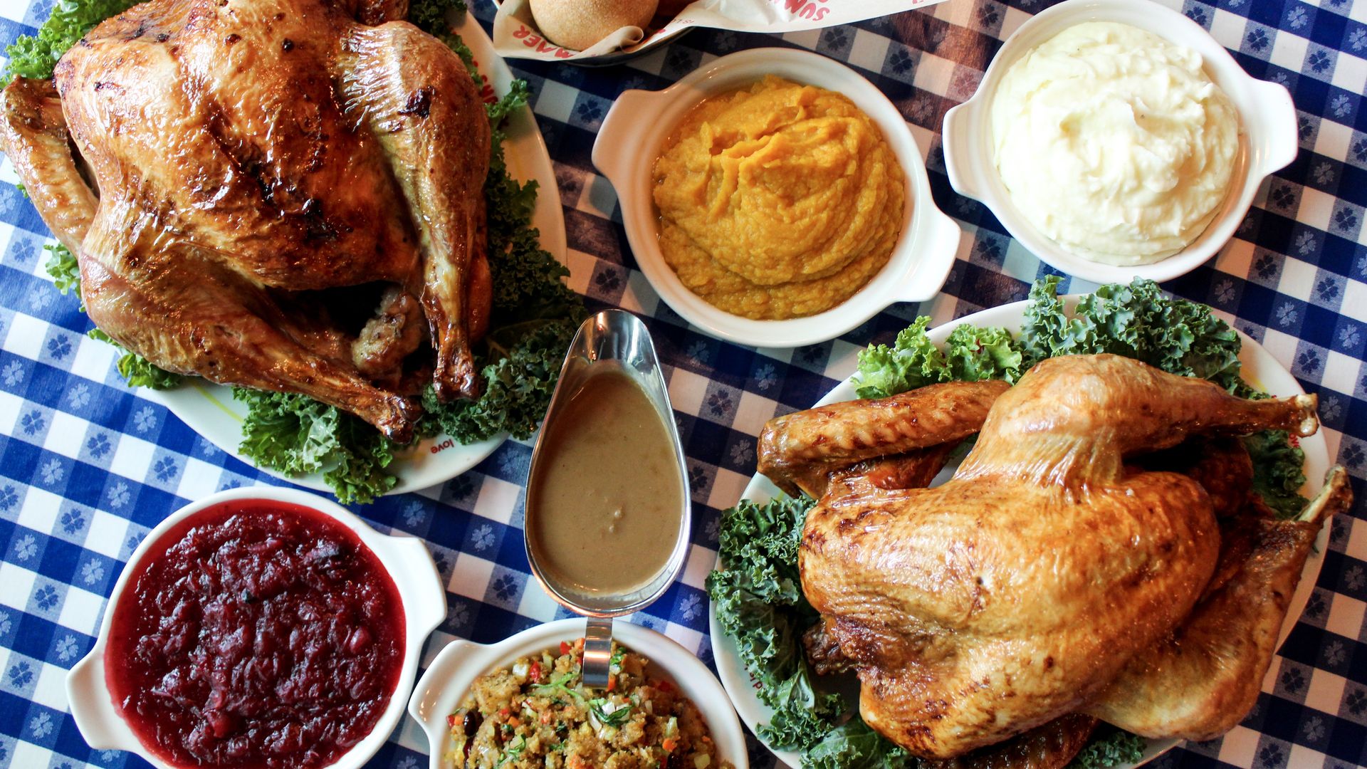 A close up of a Thanksgiving feast  from Summer Shack laid over a blue and white table setting: There are two whole turkeys, gravy, cranberry sauce, butternut squash, mashed potatoes and rolls.