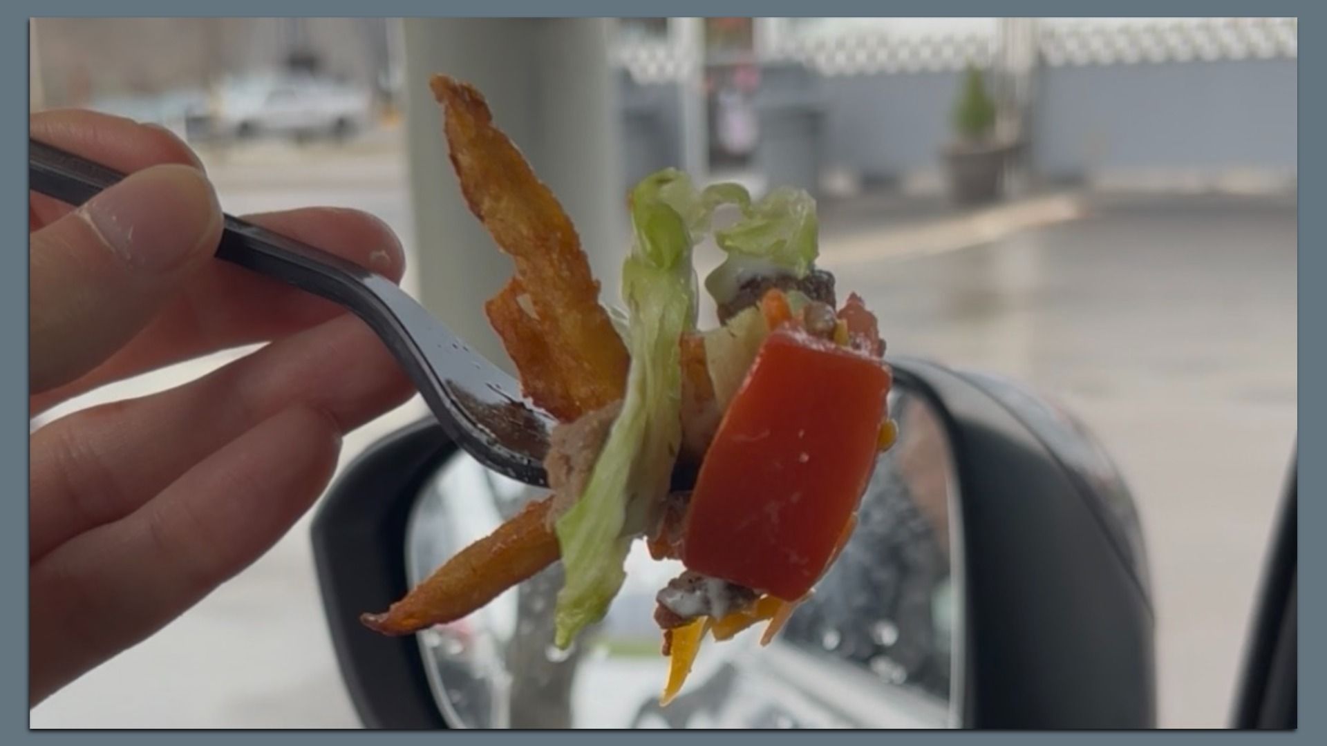 Close-up of a hand holding a forkful of salad with lettuce, red bell pepper, carrot strips, and dressing, held up near a car window outdoors.
