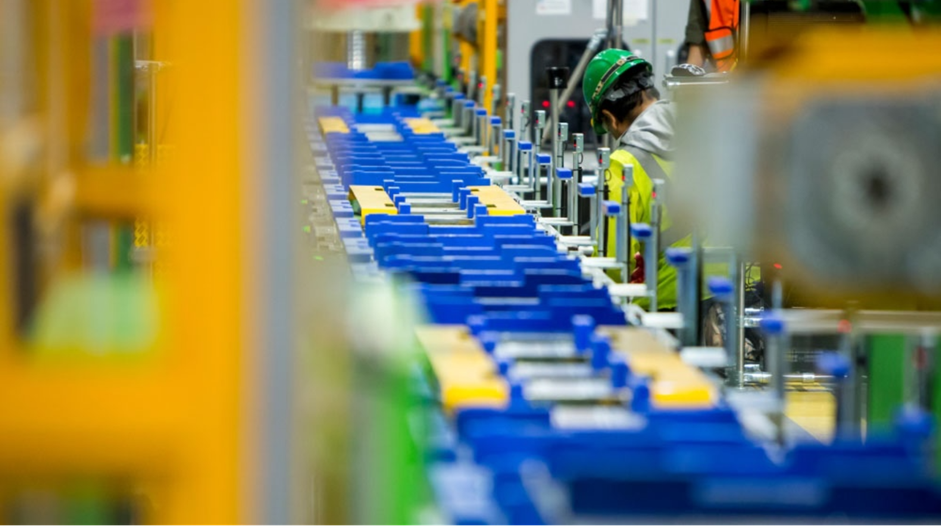 Worker wearing a green hard hat and yellow safety jacket inspecting blue and yellow items on a long industrial conveyor belt in a factory setting.