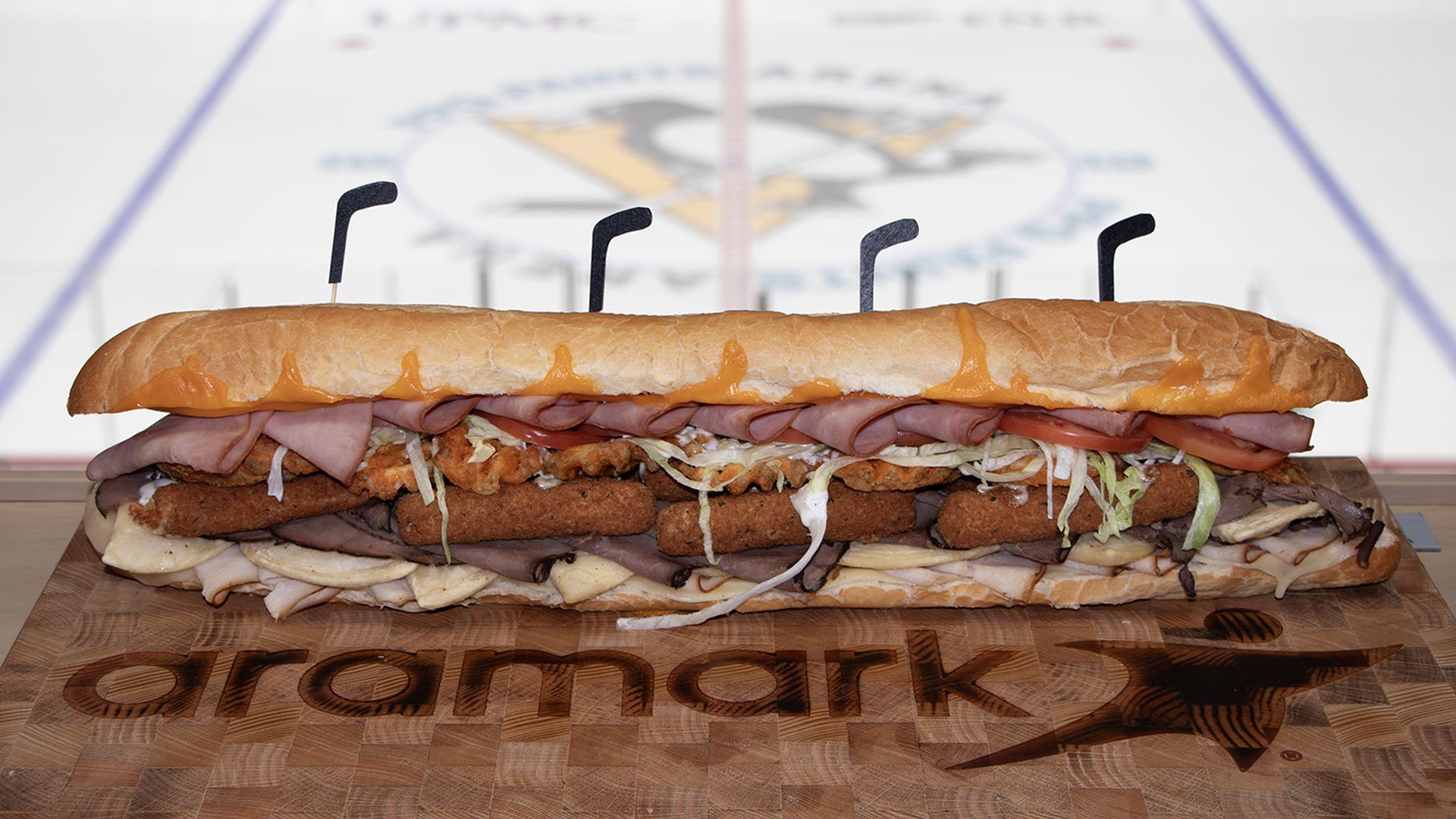 A giant submarine sandwich on a wooden cutting board, layered with ham, fried patties, shredded lettuce, onions, and tomatoes, secured by four skewers; blurred hockey rink in the background.