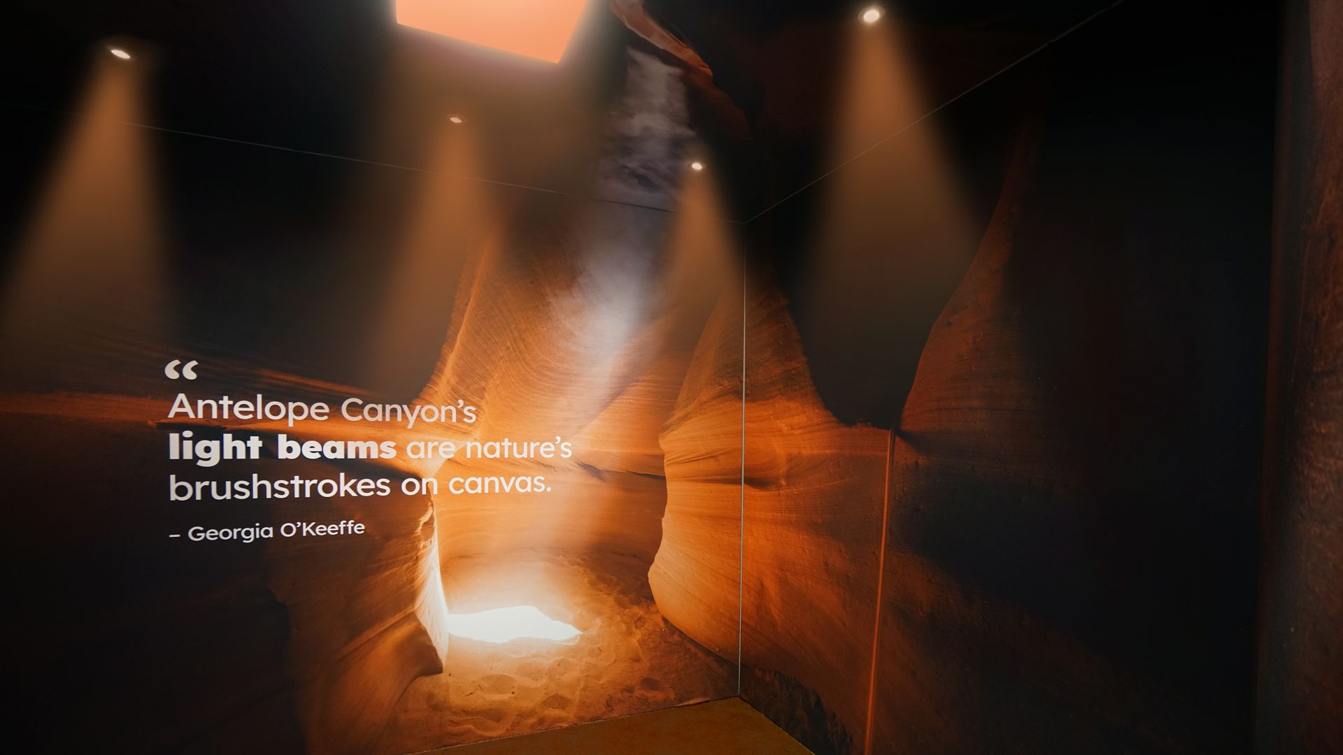 Exhibit wall bathed in warm orange spotlights, showing canyon textures. A large quote reads: "Antelope Canyon's light beams are nature's brushstrokes on canvas." — Georgia O'Keeffe.