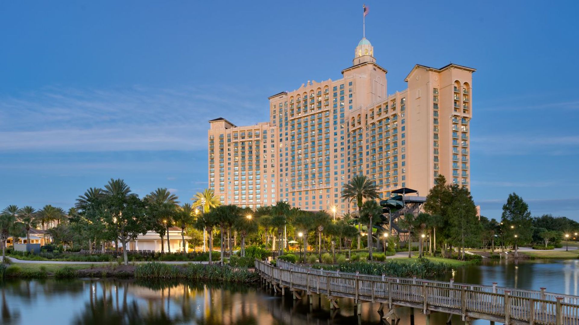 Beige hotel with twin wings and a central tower rises beside a calm lake. Palm trees line a lit promenade and a wooden boardwalk crosses the water under a blue evening sky.