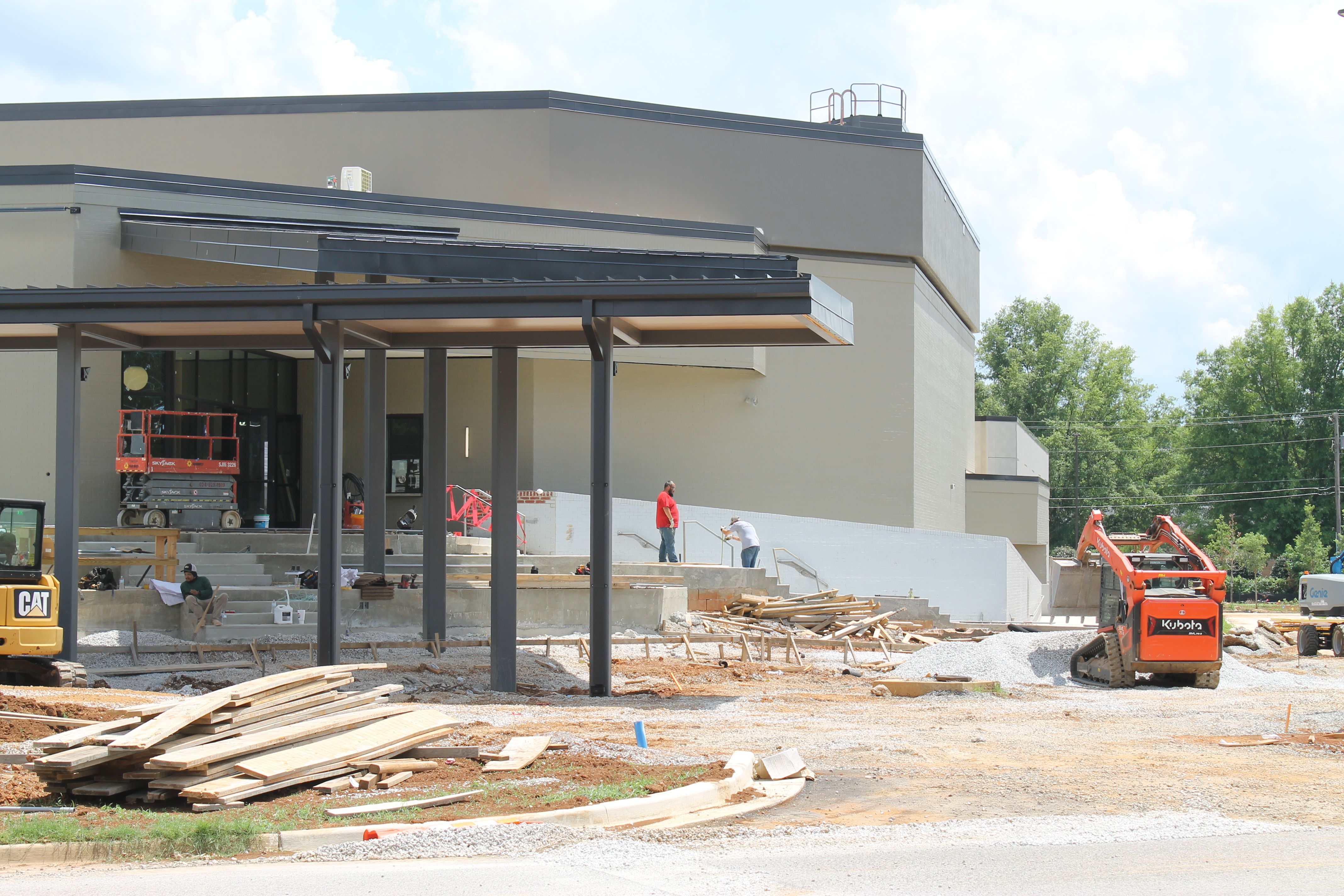 Construction site featuring a beige building under development with black metal framework and overhangs. Lumber piles, construction vehicles (CAT, Kubota), and workers in red shirts; blue sky with clouds.
