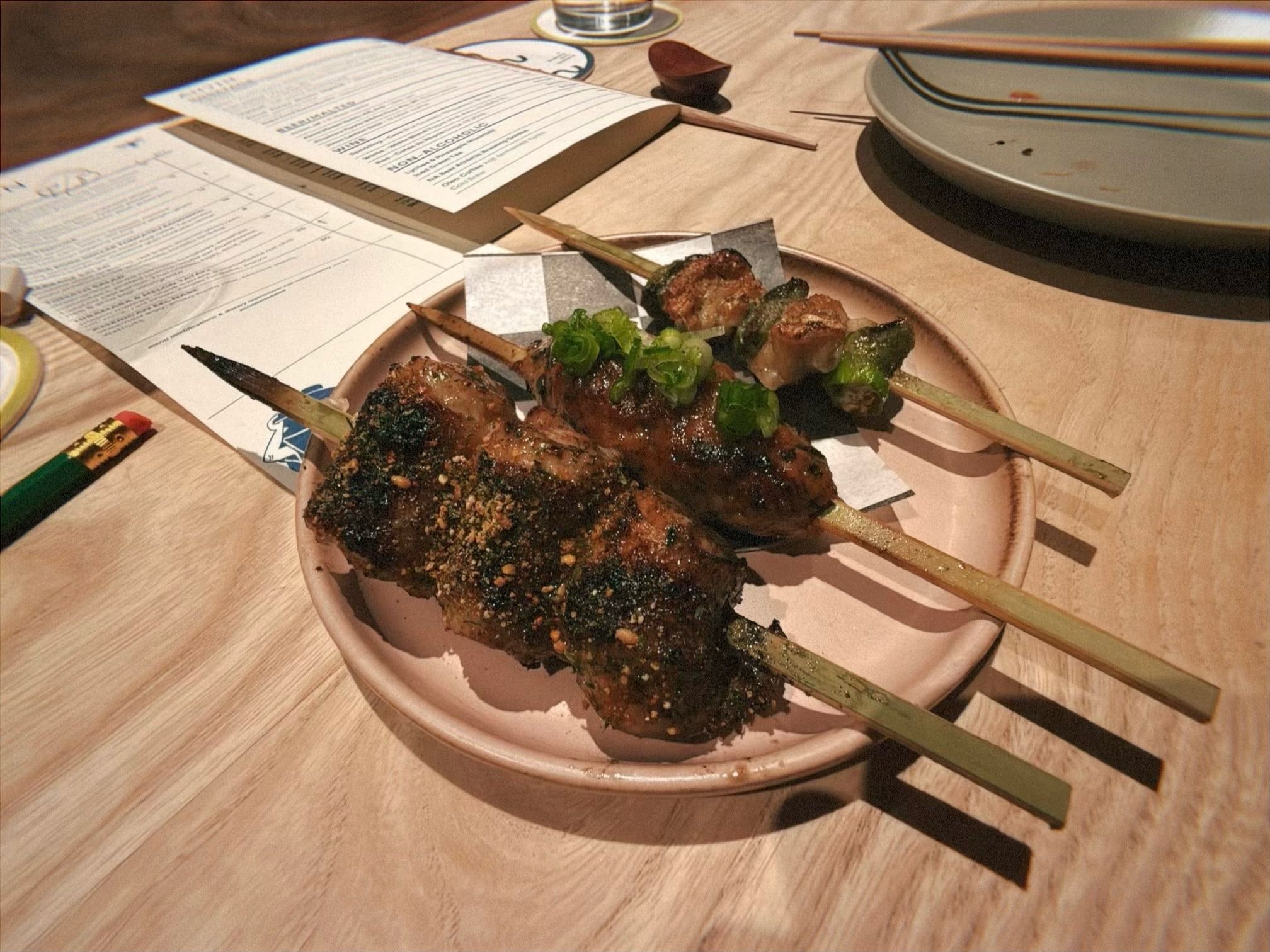 Three grilled skewers with meat and green garnishes on a beige plate, placed on a light wooden table with menus, chopsticks, and a coaster nearby.