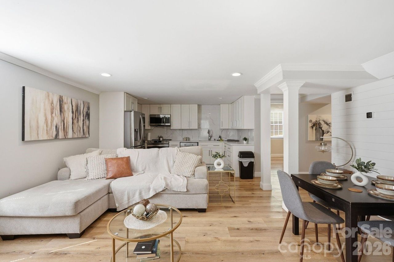 Open-plan living area with a light gray L-shaped sofa, white throws and a peach pillow, round glass coffee table, and abstract wall art. In the background is a white kitchen; to the right, a dark dining table with gray chairs on light wood floors.