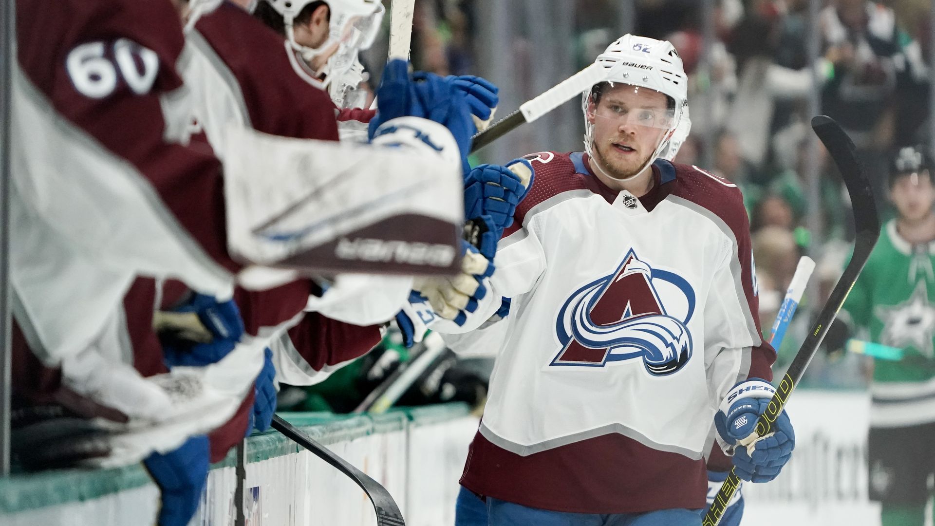 A man in a helmet and large jersey walks past teammates behind a small wall.