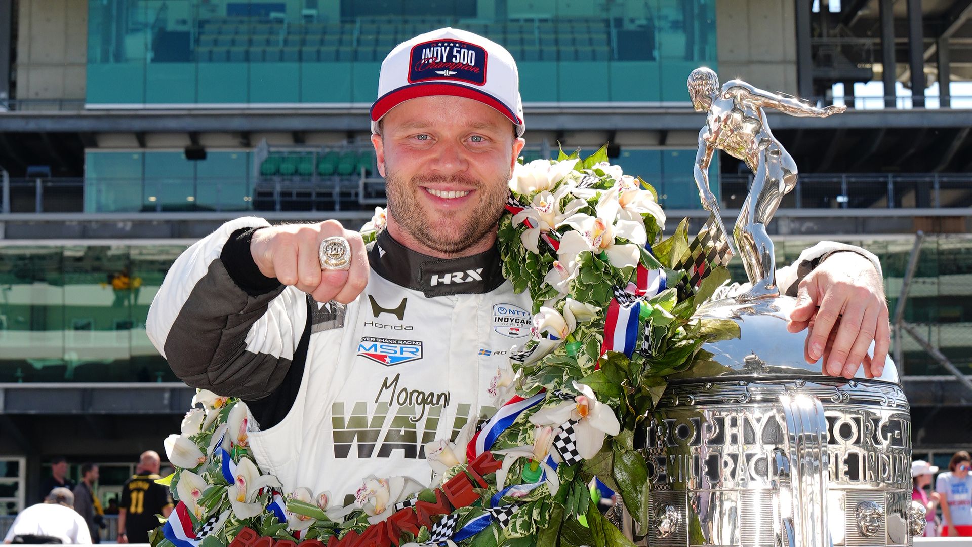 Race car driver in a white racing suit and cap reading "INDY 500", smiling amid a flower garland, posing with a tall silver trophy topped by a figure, with a glass stadium backdrop.