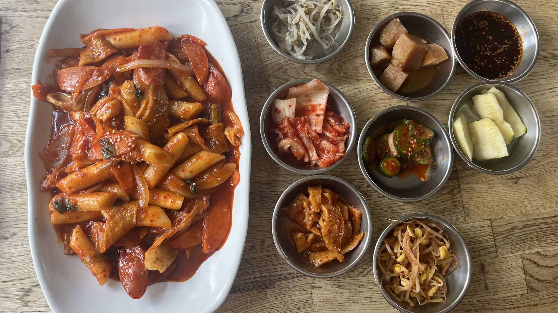 Plate of spicy Korean rice cakes (tteokbokki) in red sauce with onions and fish cakes, accompanied by seven small bowls of Korean side dishes including kimchi, pickled cucumbers, and dipping sauce on a wood table.