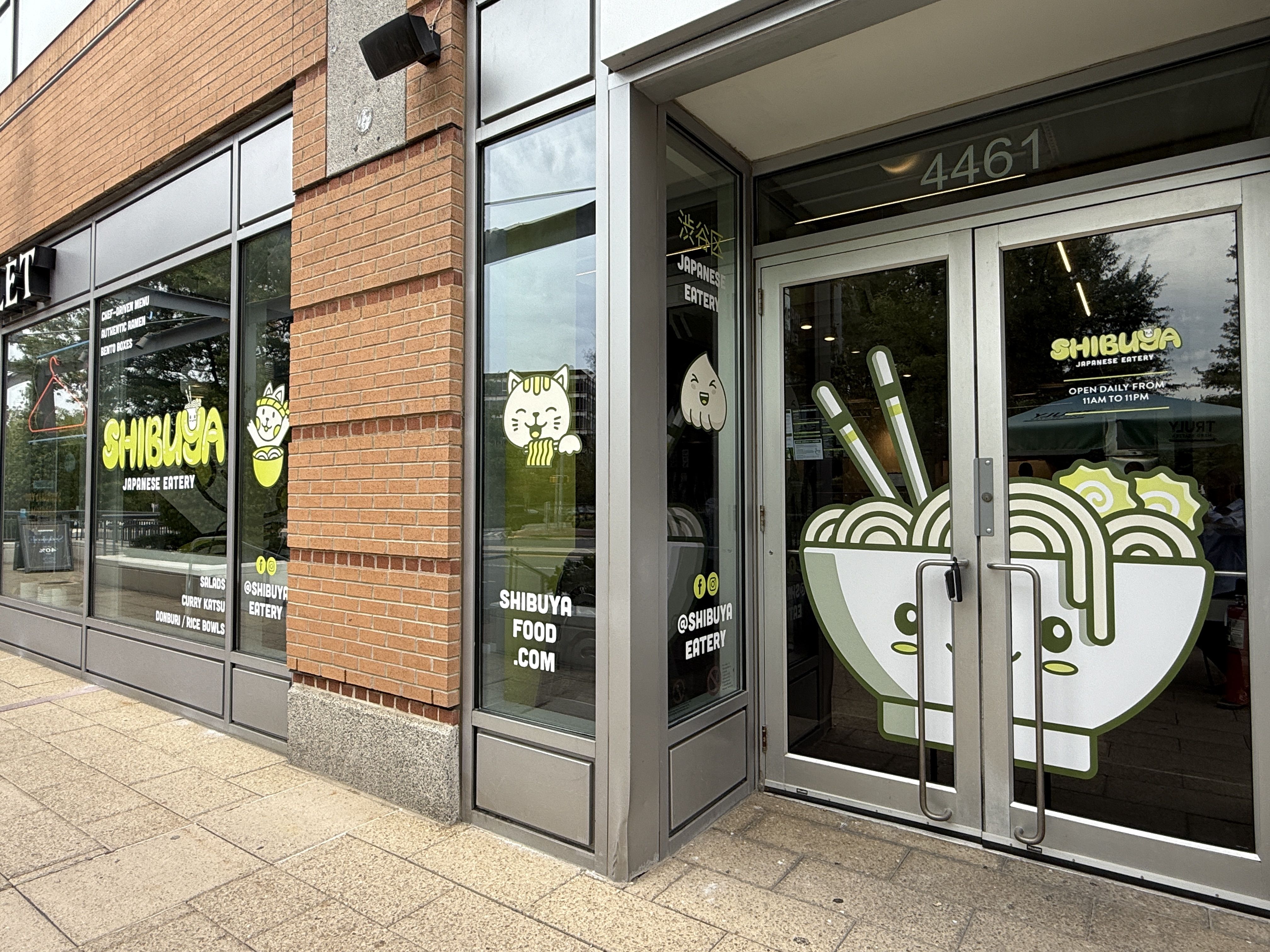 Entrance of Shibuya Japanese Eatery with cartoon ramen bowl and cat graphics on glass doors and windows, brick and concrete building exterior, address 4461.