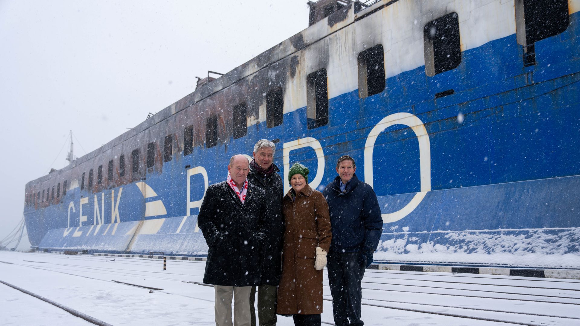 Four people dressed in winter coats stand in the snow in front of a large blue and white ship with visible rust and blackened areas, snow falling around them.