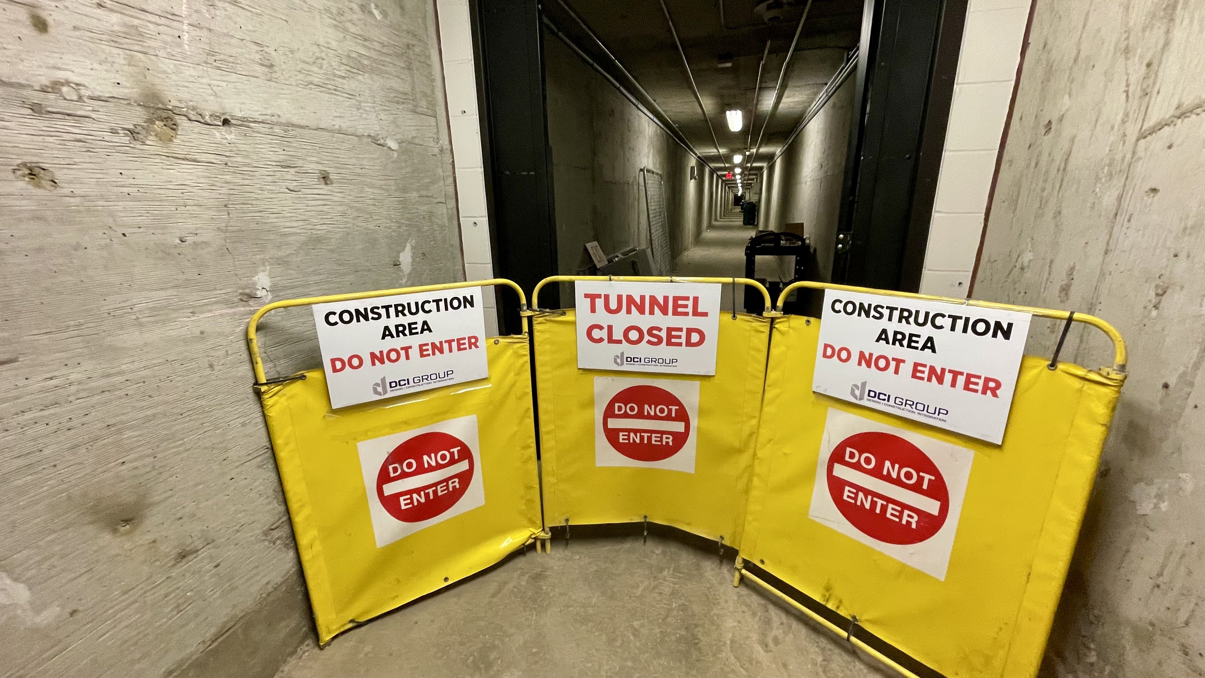 Yellow barriers block a concrete tunnel entrance, with signs reading "Tunnel Closed," "Construction Area," and "Do Not Enter" in red and black text. The tunnel extends darkly behind.
