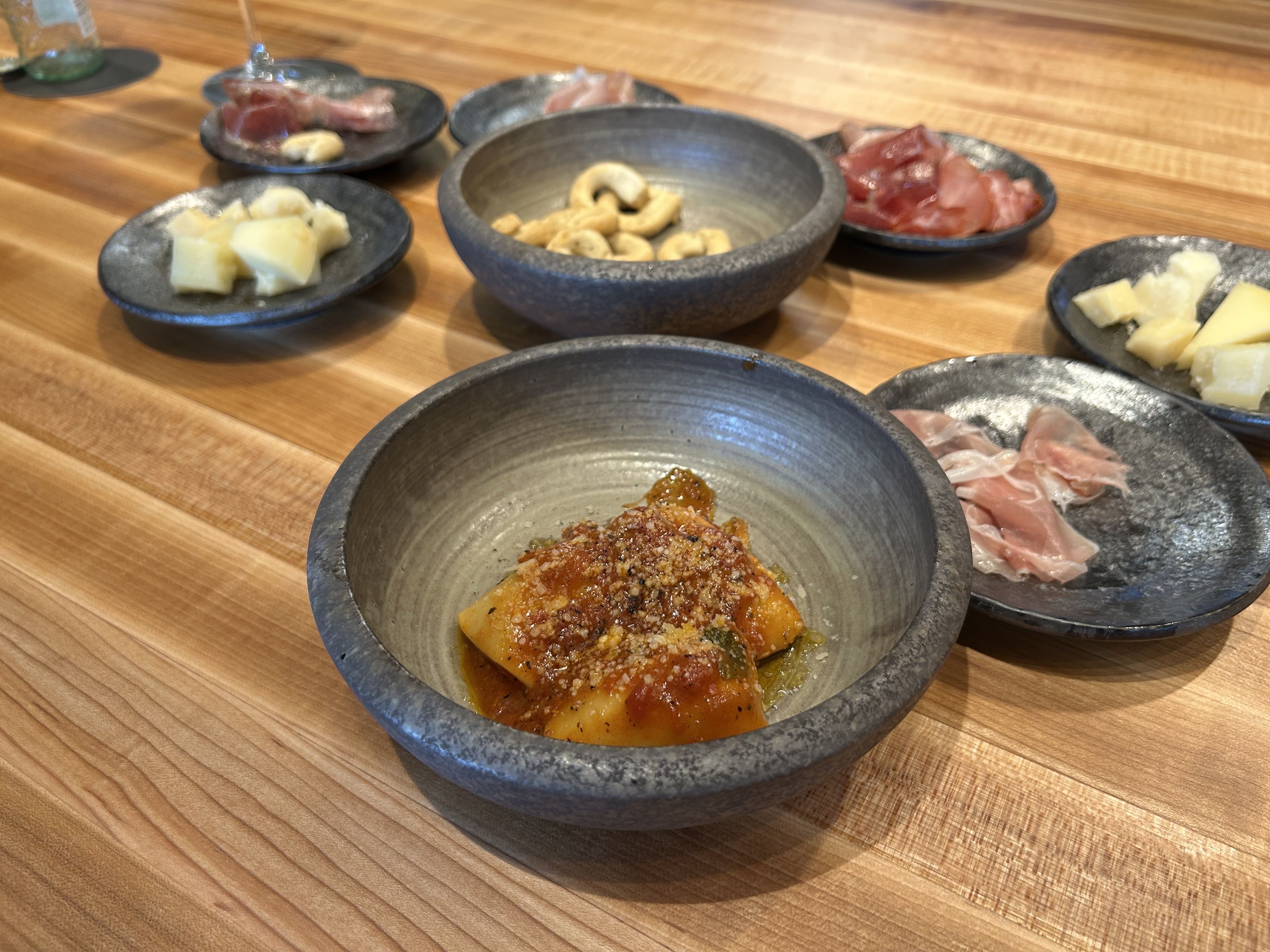 Several small stone bowls on a wooden table containing various foods including cheese cubes, cured meats, cashews, and a bowl with orange pasta topped with tomato sauce and grated cheese.