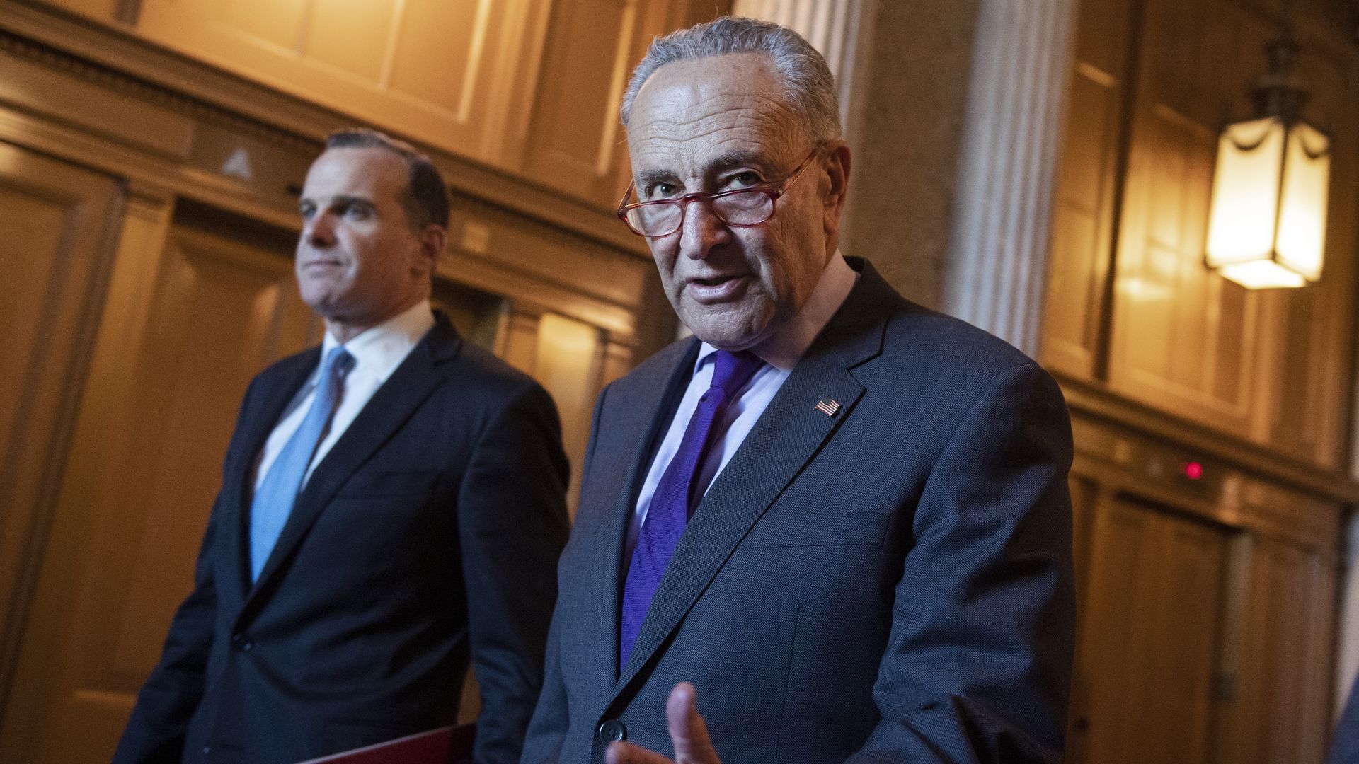 In this image, Schumer talks and gestures while wearing a suit and walking down a hallway.