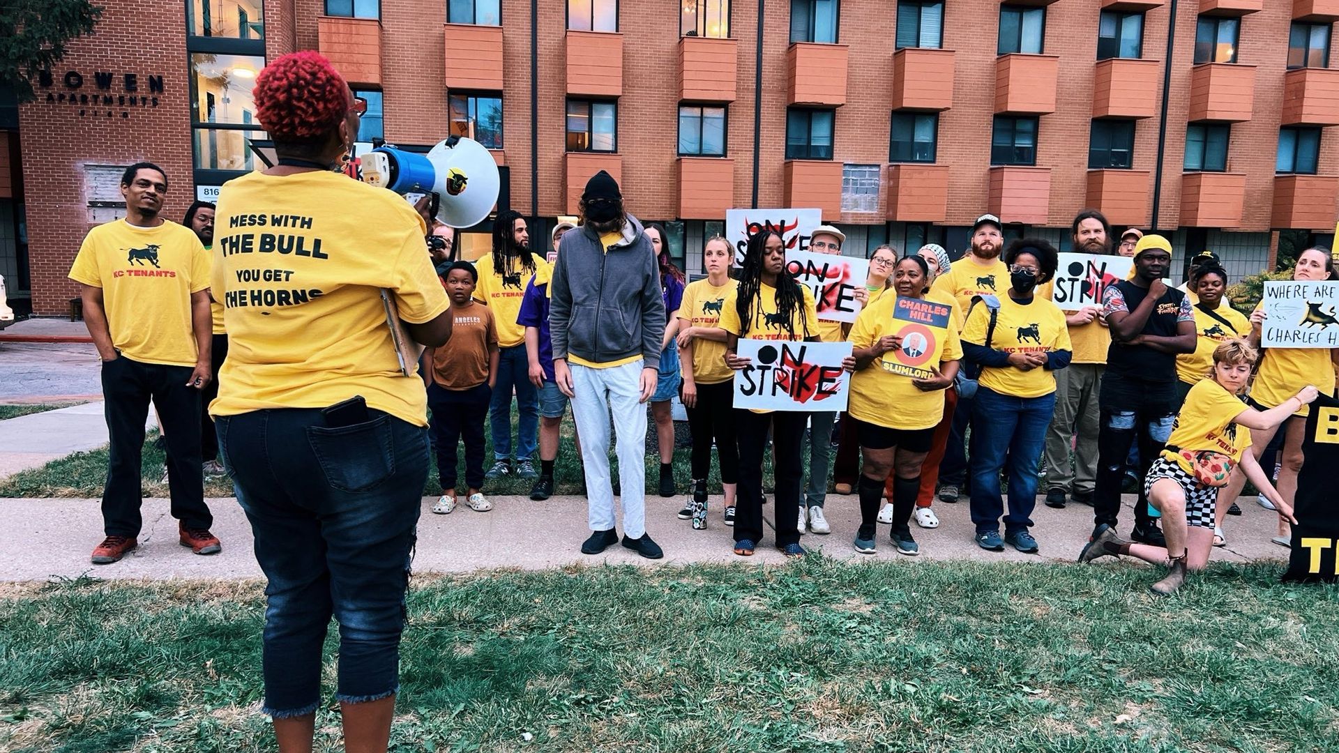 Group of people wearing yellow KC Tenants shirts protest outside an apartment building holding signs reading "On Strike" and asking about Charles Hill, with one person speaking on a megaphone.