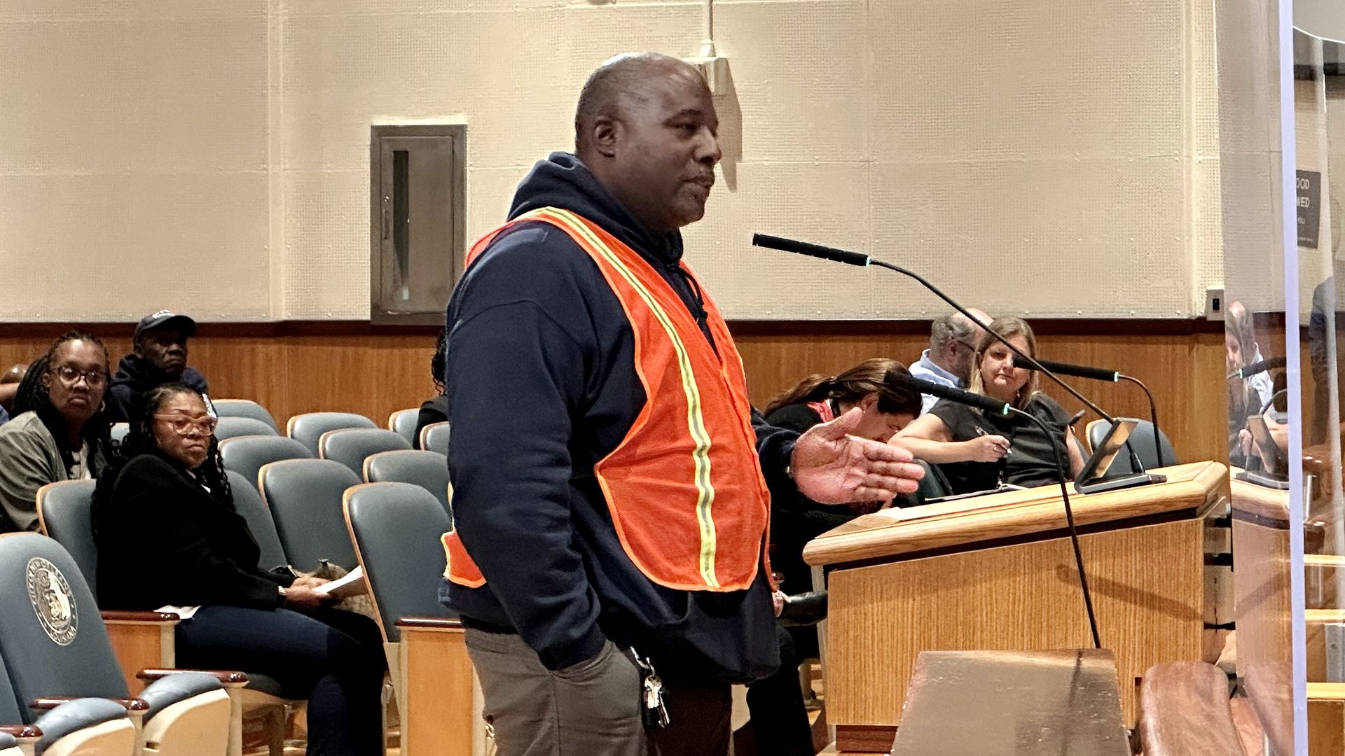 Man wearing an orange safety vest over a dark hoodie speaks at a wooden podium with a microphone in a council chamber as attendees sit and listen.