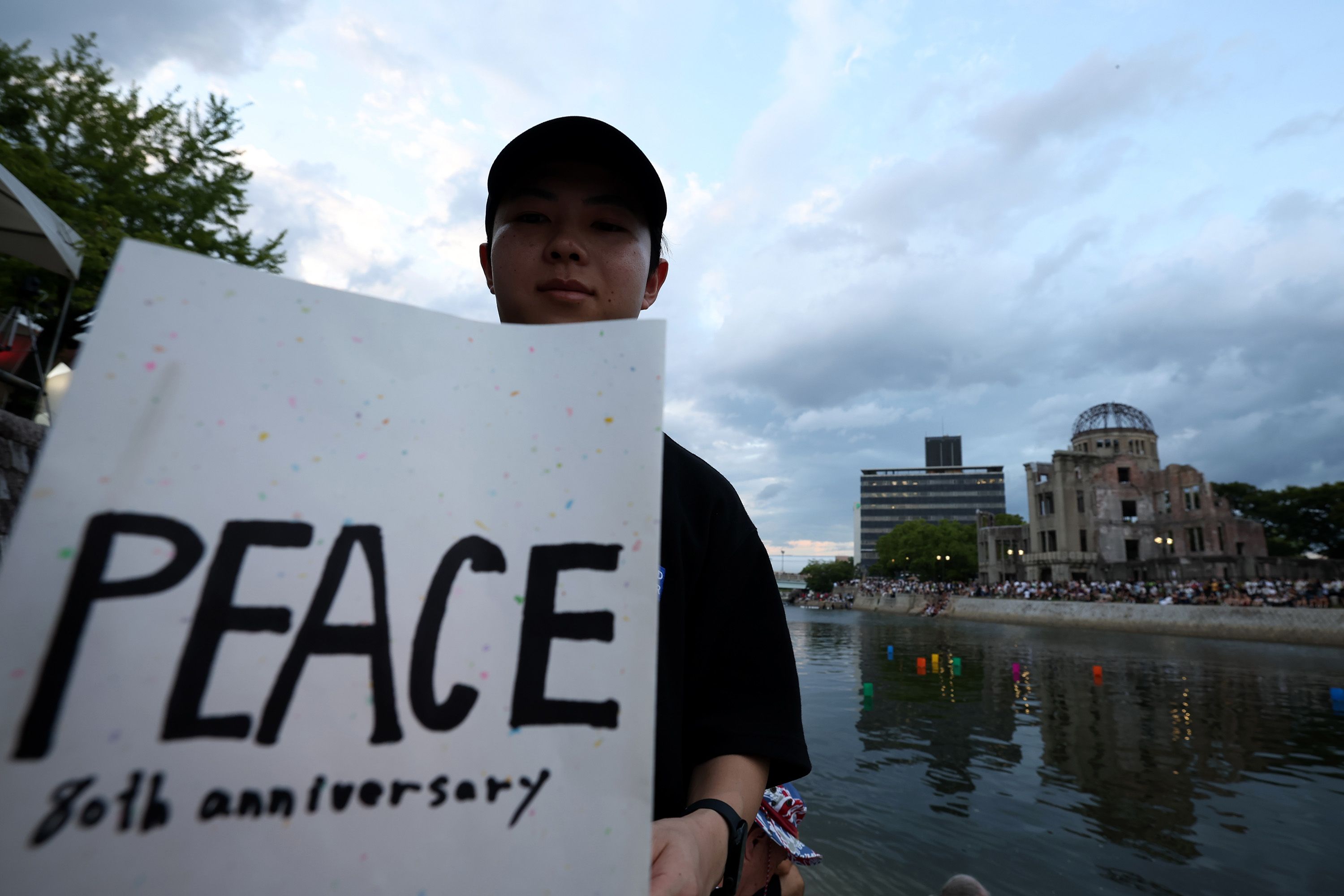 Person holding a sign reading "PEACE 80th anniversary" near the river with the atomic bomb dome in Hiroshima and a crowd gathered at dusk under a cloudy sky.