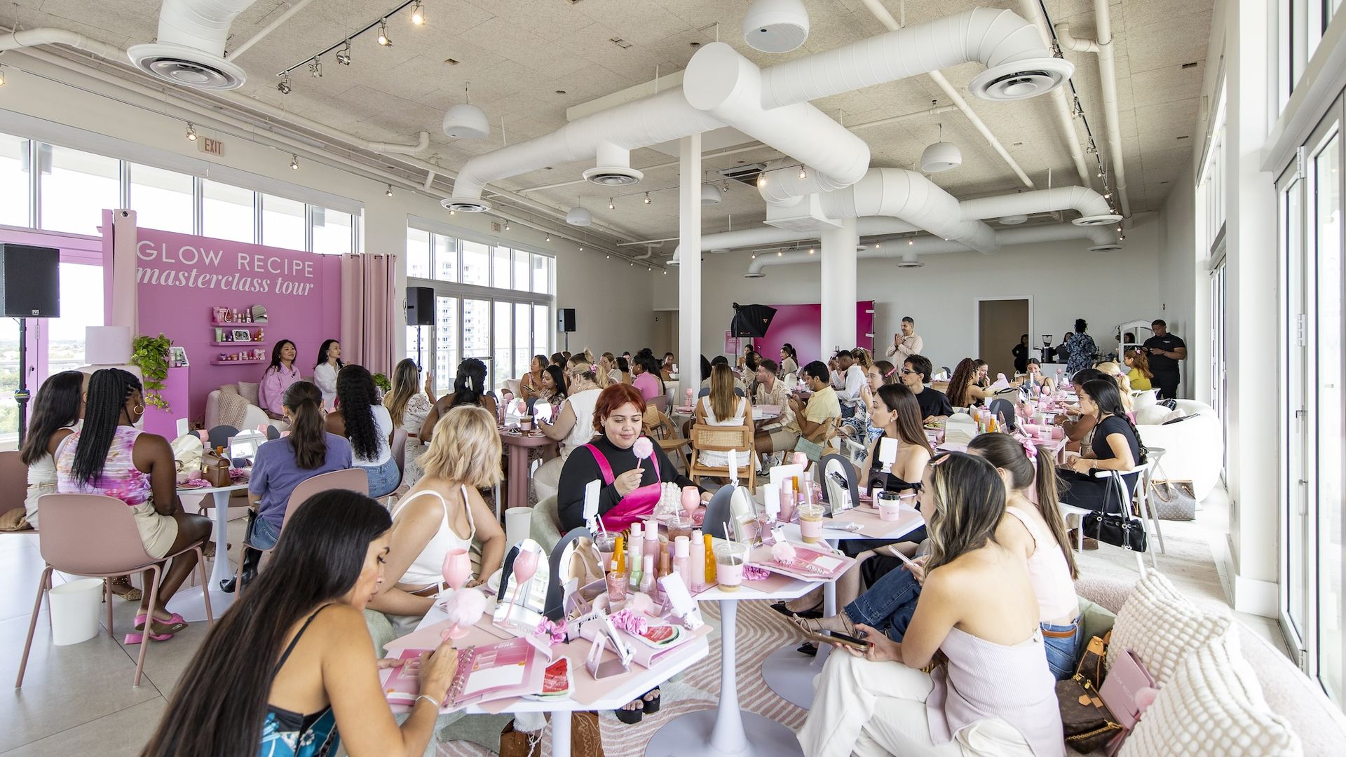 People sitting at white tables with pink bottles on the tables while two women are on a stage with a pink banner behind them.