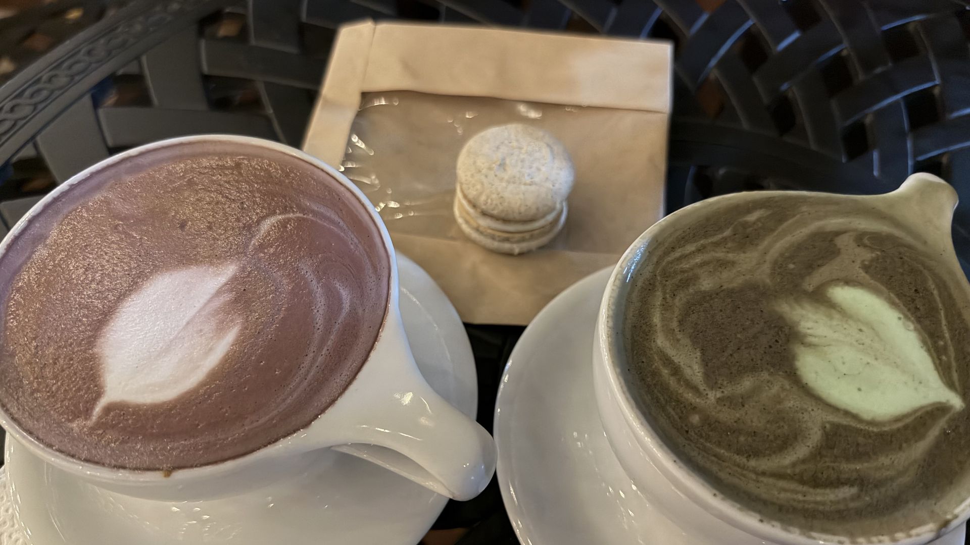 Top-down view of two cups on white saucers with foamy drinks. Left: pink-lavender latte; right: greenish matcha latte. A small stack of macarons sits on a napkin behind them.