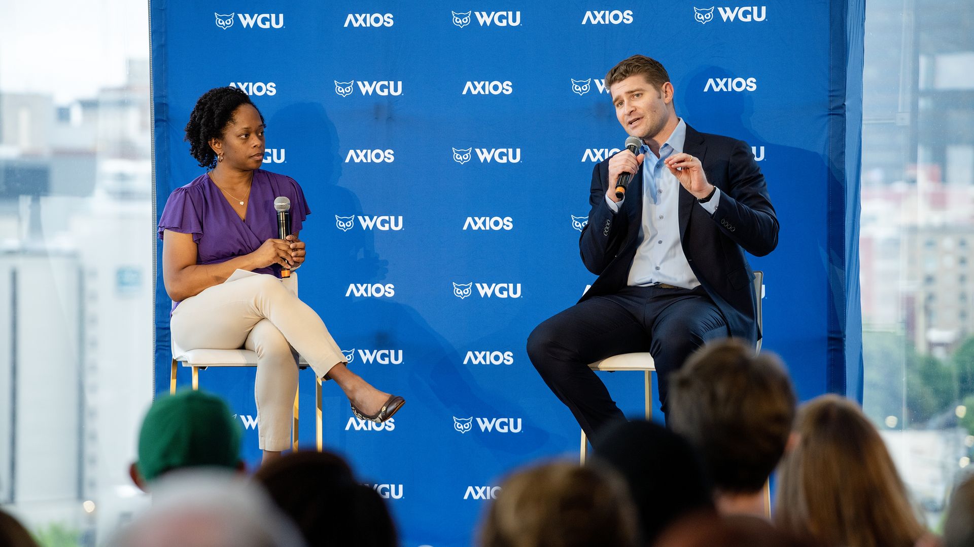 Two panelists on a stage with a blue backdrop featuring WGU and Axios logos; a woman in purple blouse holds a microphone while a man in a dark suit speaks into a microphone, addressing audience.