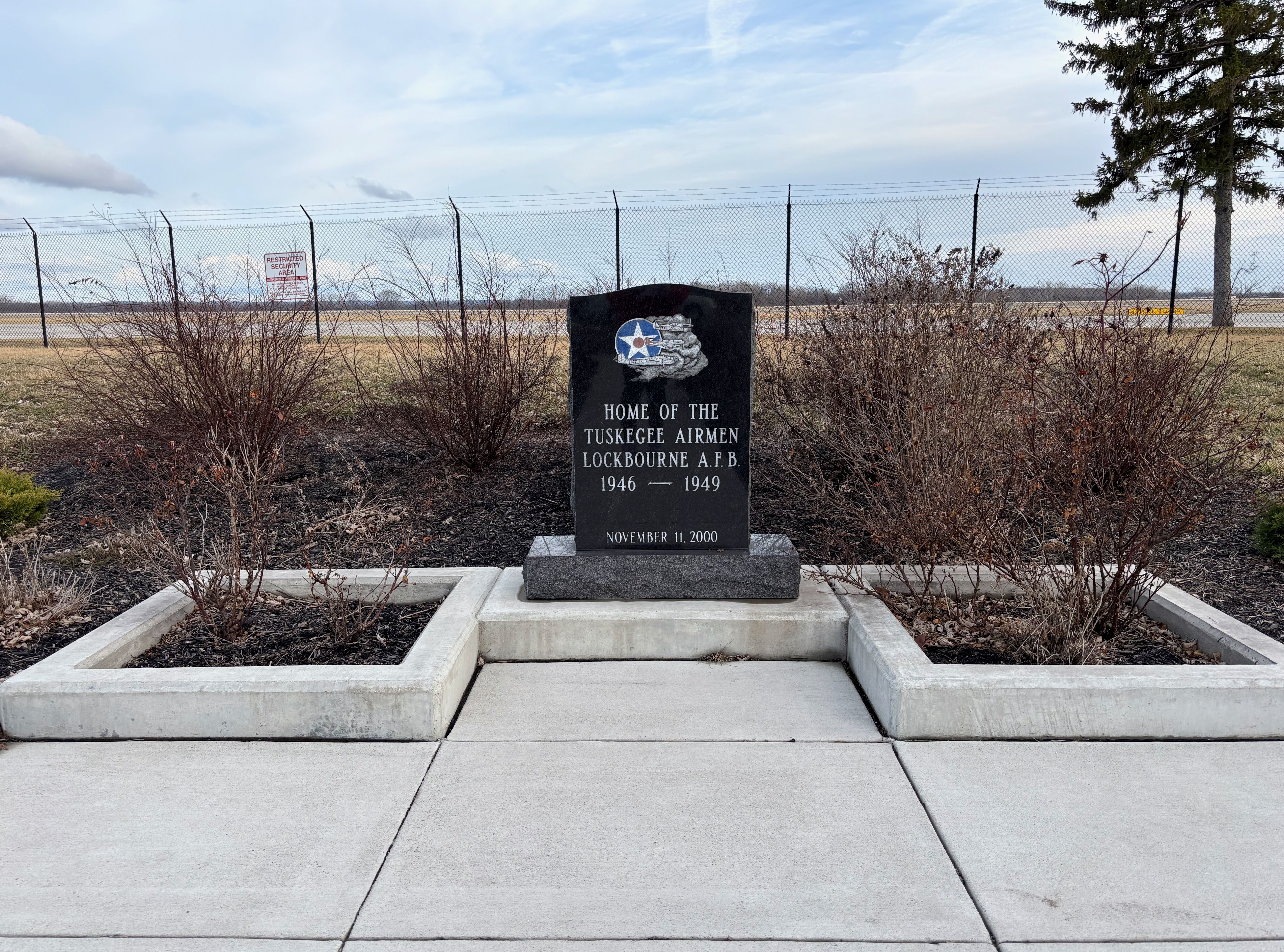Black memorial stone with Air Force symbol and inscription honoring Tuskegee Airmen at Lockbourne A.F.B. 1946-1949, surrounded by bare shrubs and set on a concrete platform against a fence backdrop.