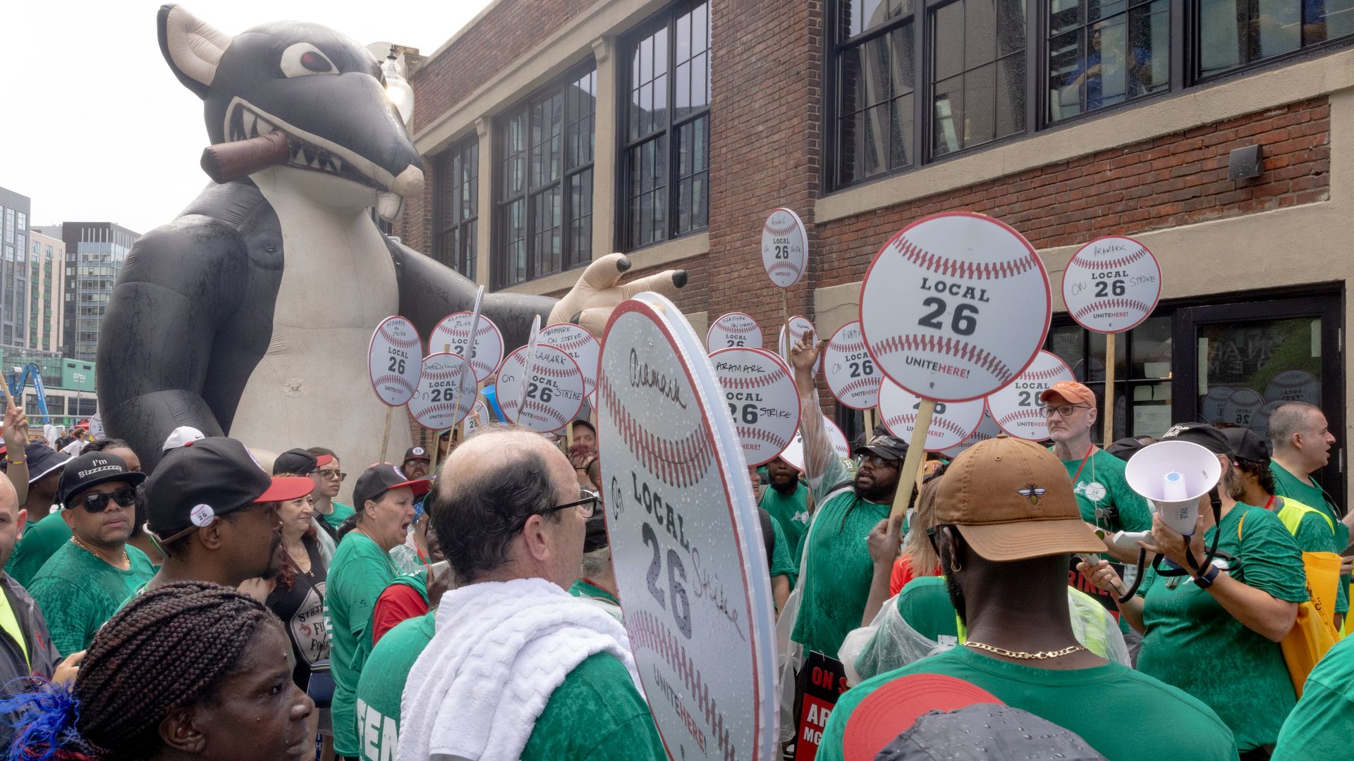 Crowd of people wearing green shirts holding baseball-shaped signs reading "Local 26 Unite Here" during a strike outside a brick building, with a large inflatable rat holding a cigar in the background.